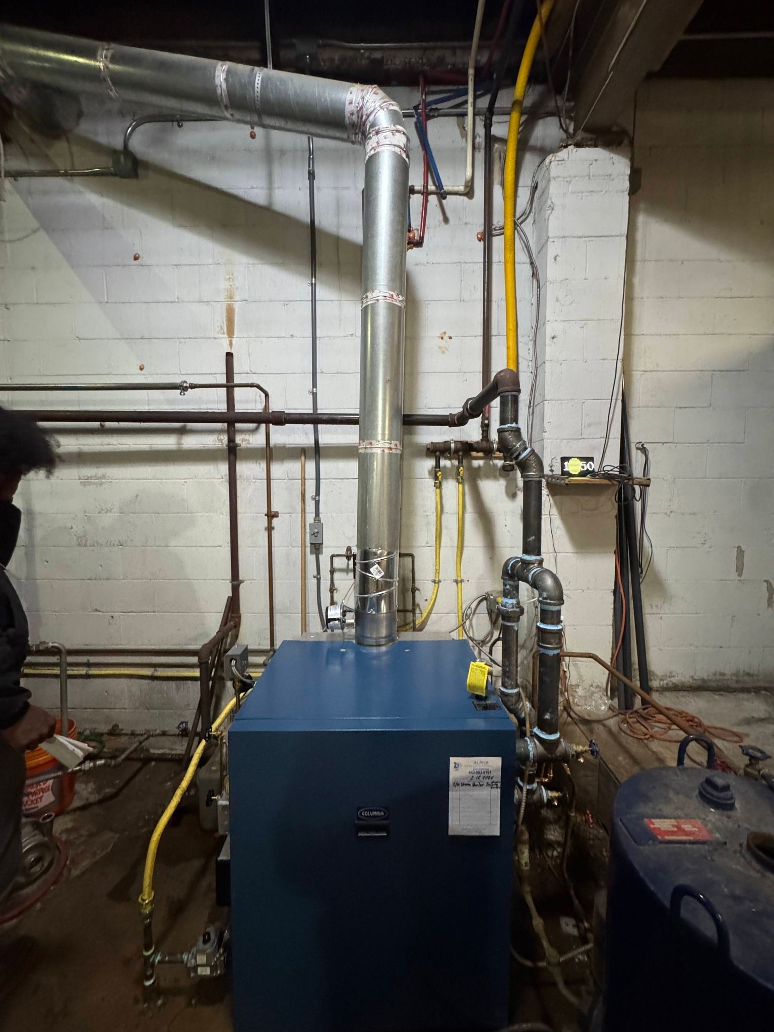 A blue commercial boiler unit in a basement, connected to silver metal exhaust ducting and yellow gas pipes.