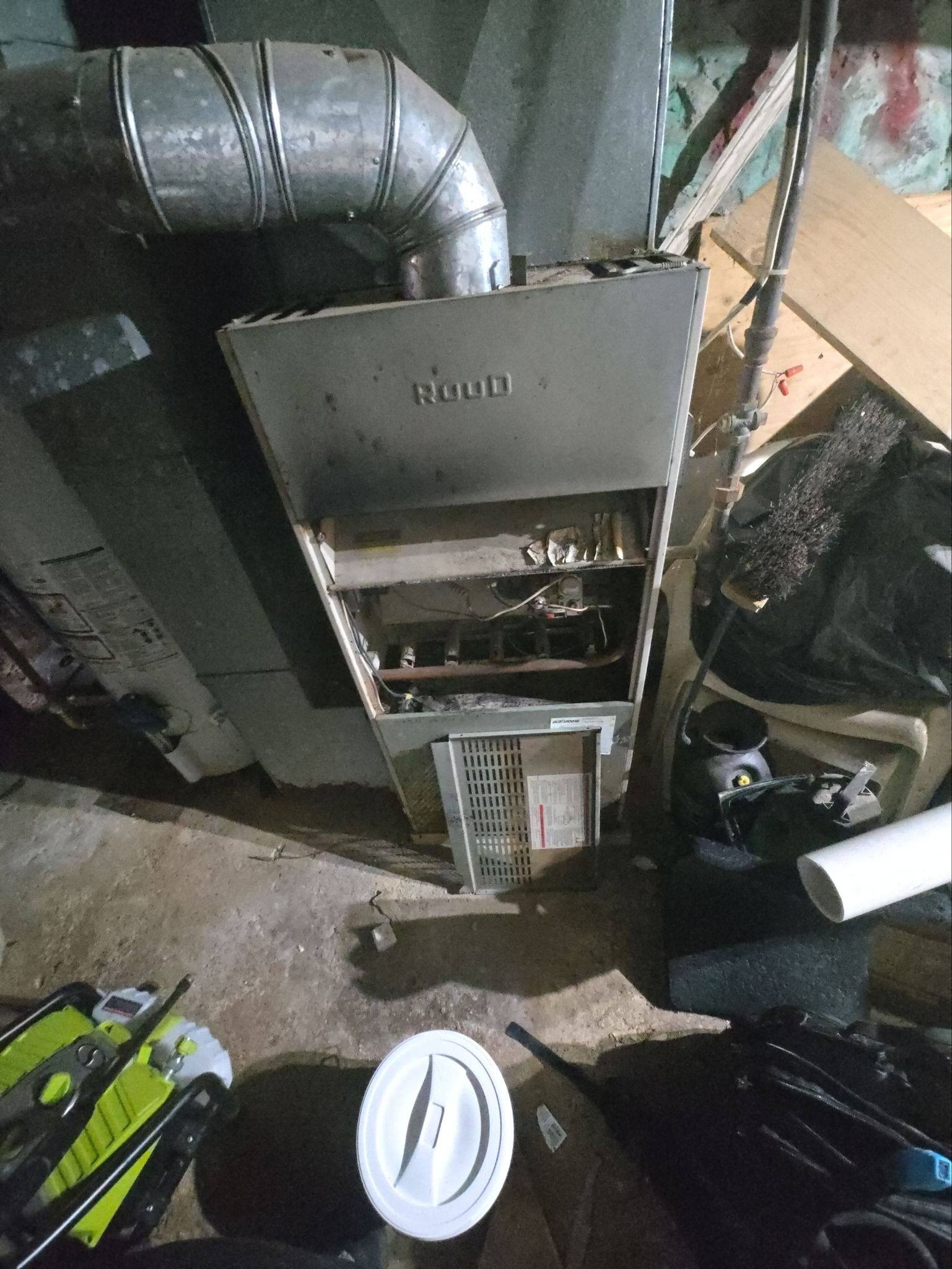 A furnace with an open access panel sits in a basement near a metal vent, some tools, and a white bucket.