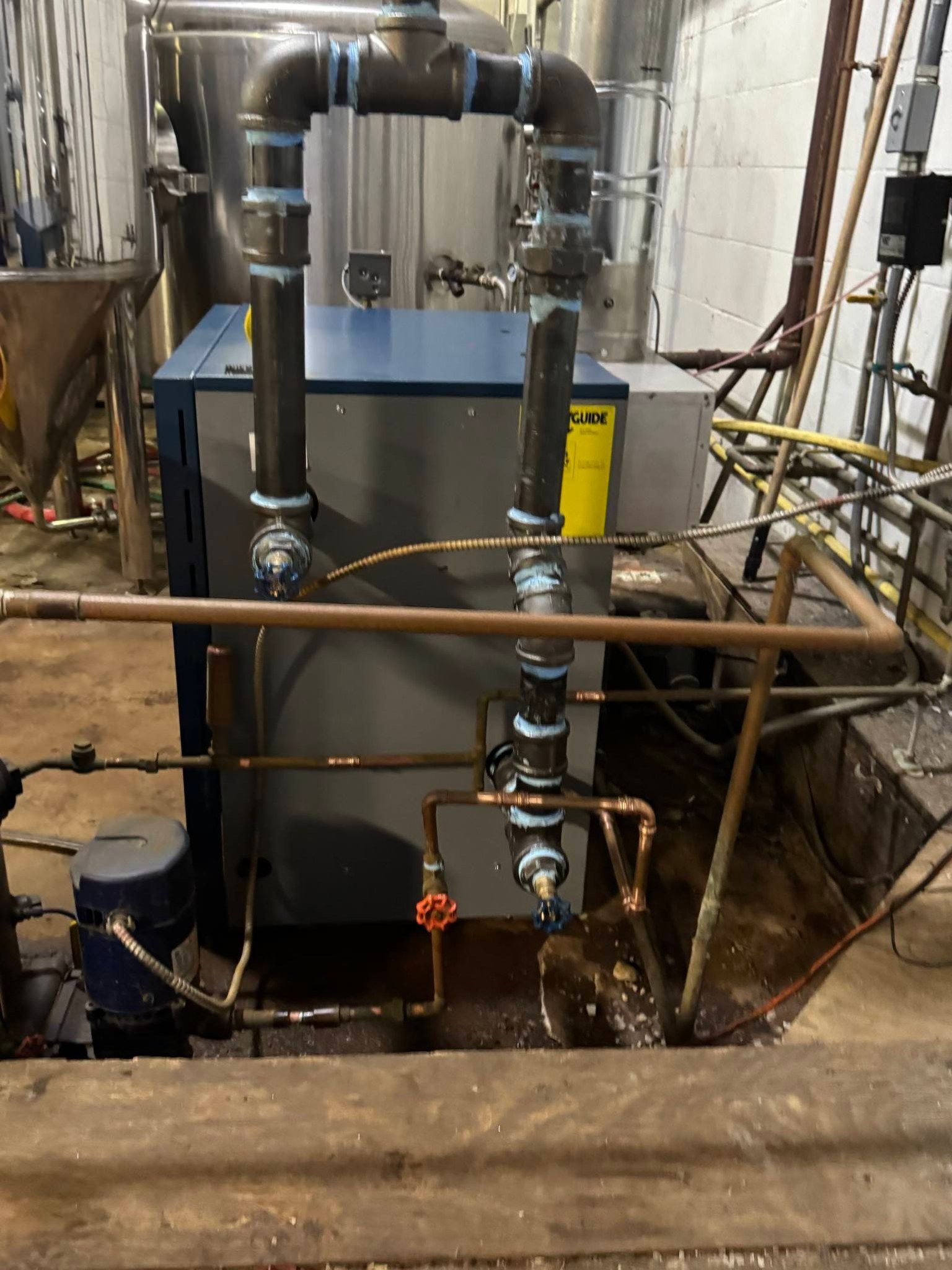 A blue and grey industrial boiler unit with complex black pipe plumbing and copper tubing in an industrial facility.