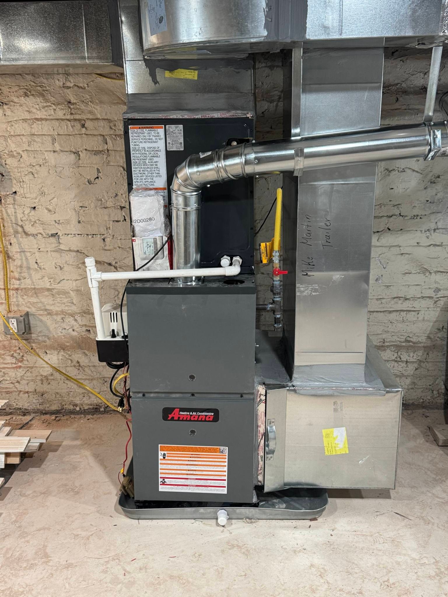 A charcoal-gray Amana gas furnace with silver metal ductwork stands against a rough concrete wall in a basement.