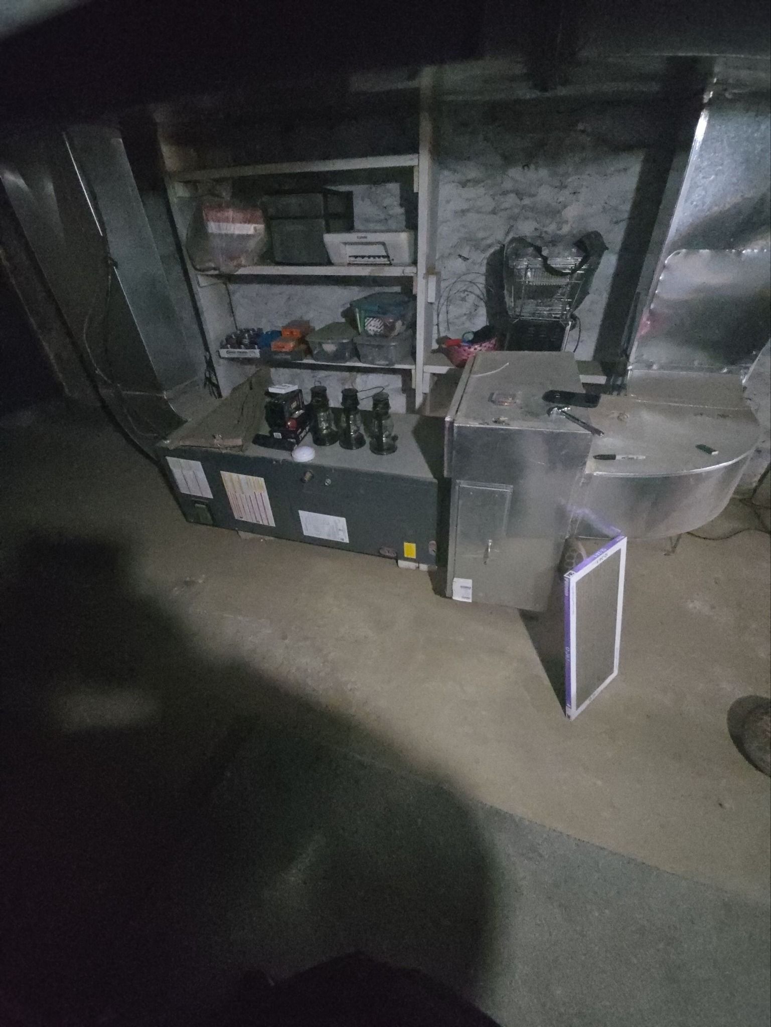 A furnace unit with exposed metal ductwork and a replacement air filter standing on a concrete basement floor.