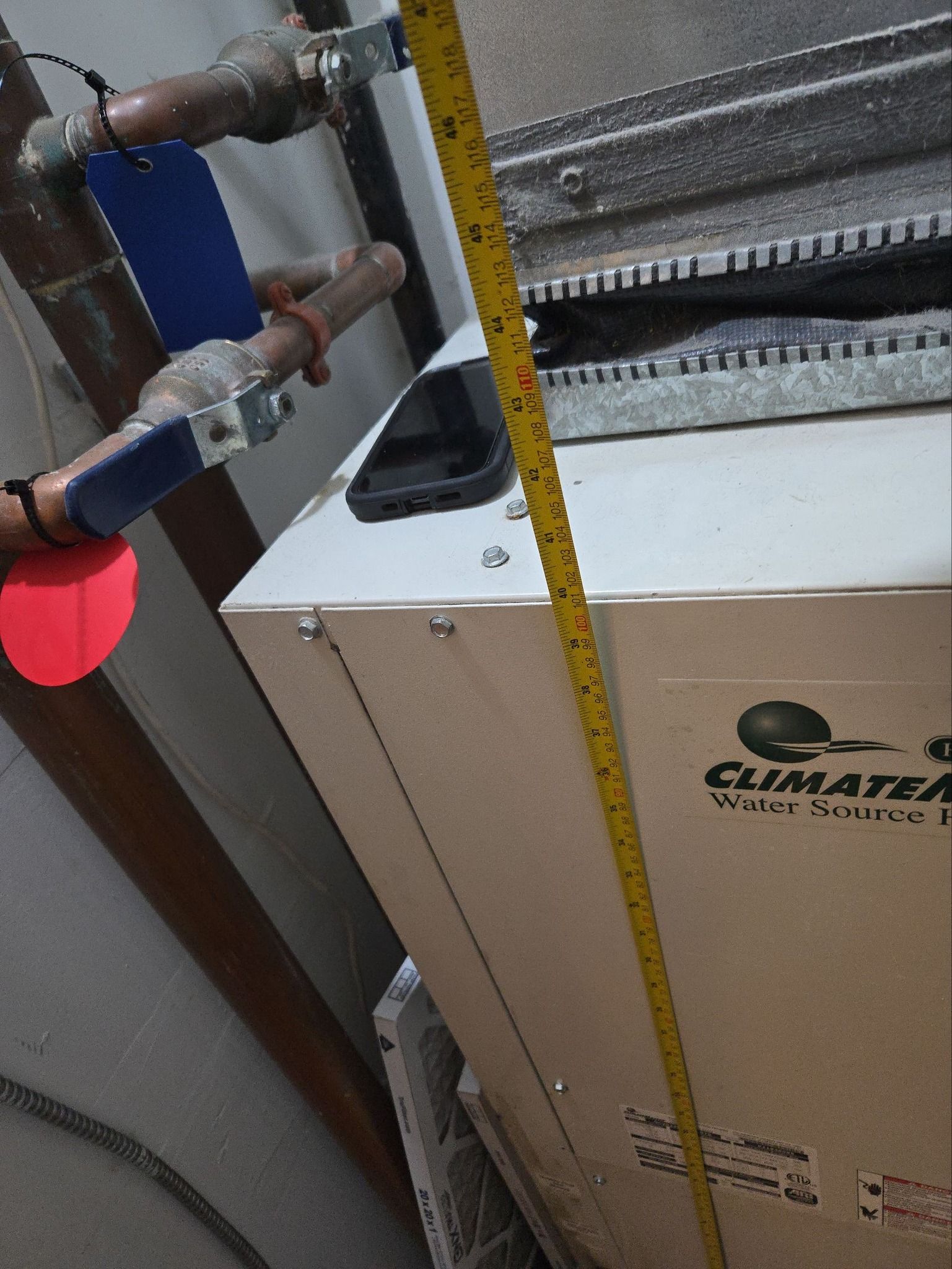 A tape measure stands vertically next to a ClimateMaster water-source heat pump unit with two copper pipes on the left.