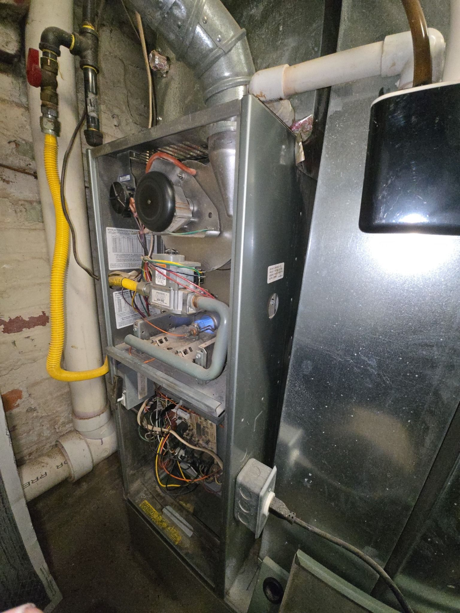A vertical metal furnace with exposed internal components, wiring, and a yellow gas line connected to overhead piping.
