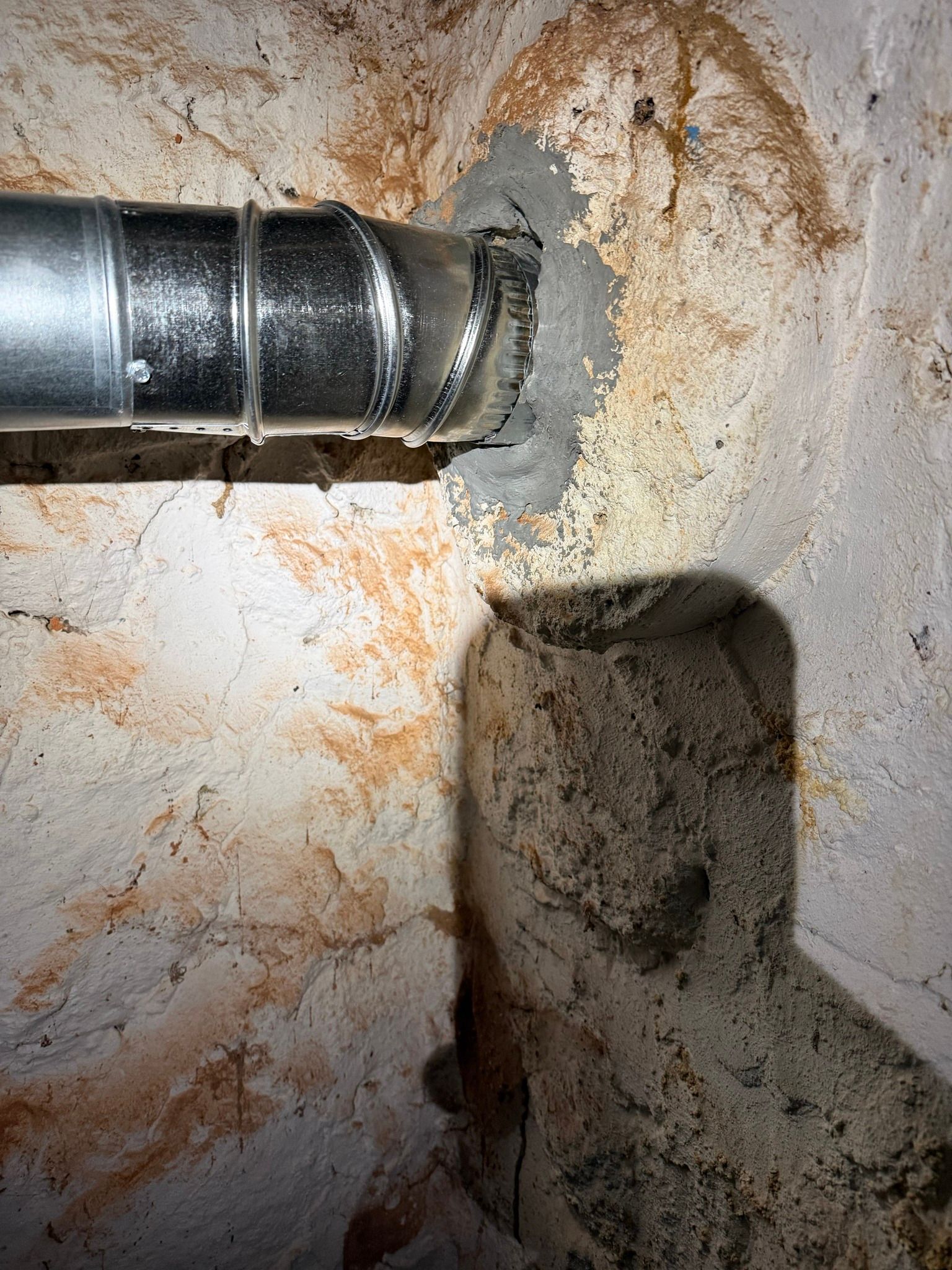 A metal pipe penetrates a rough, textured wall, sealed with grey mortar, with a shadowed recess in the wall below.