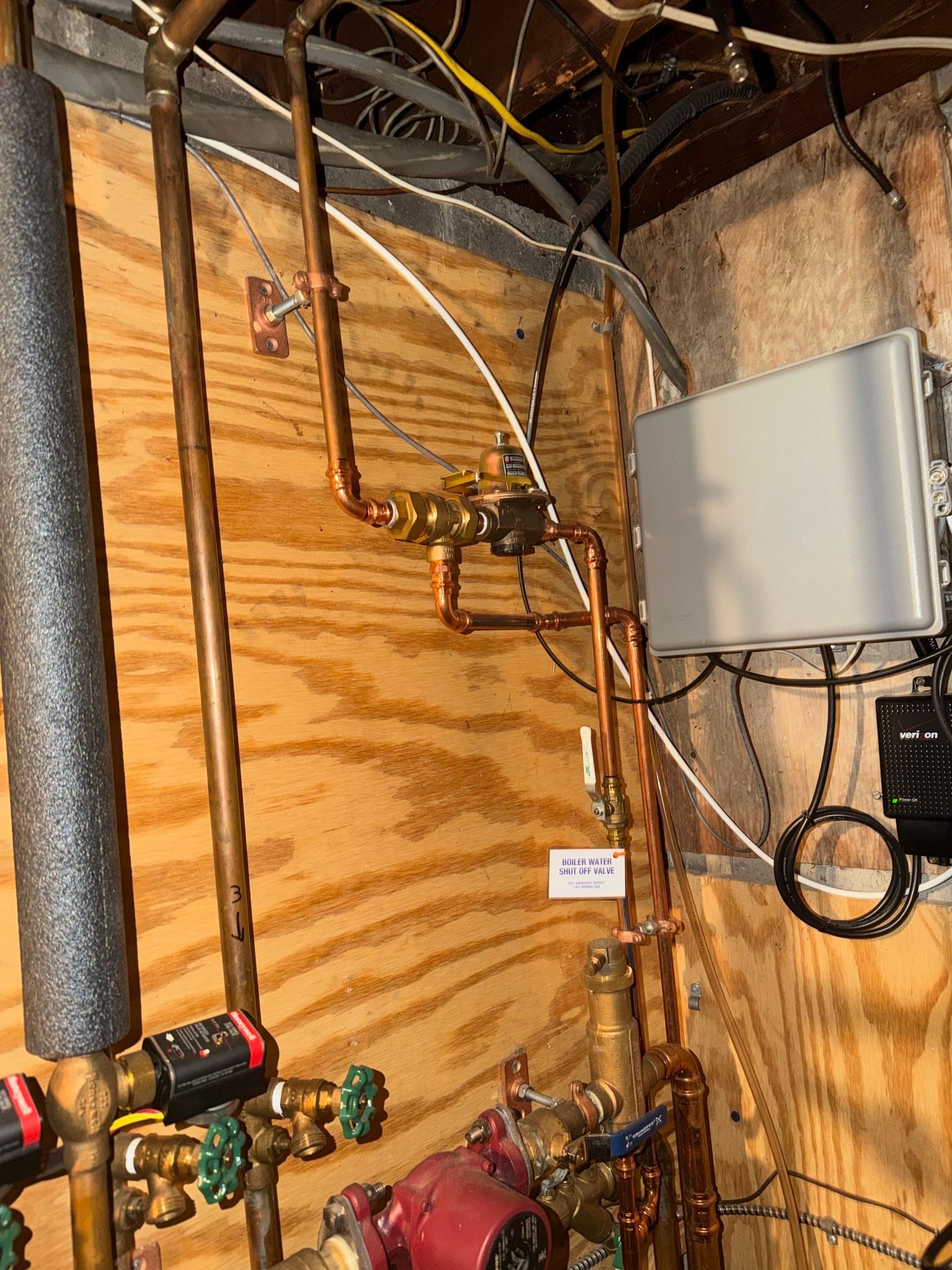 Copper plumbing pipes, valves, and a red pump mounted on a plywood wall in a utility area with an electrical box.