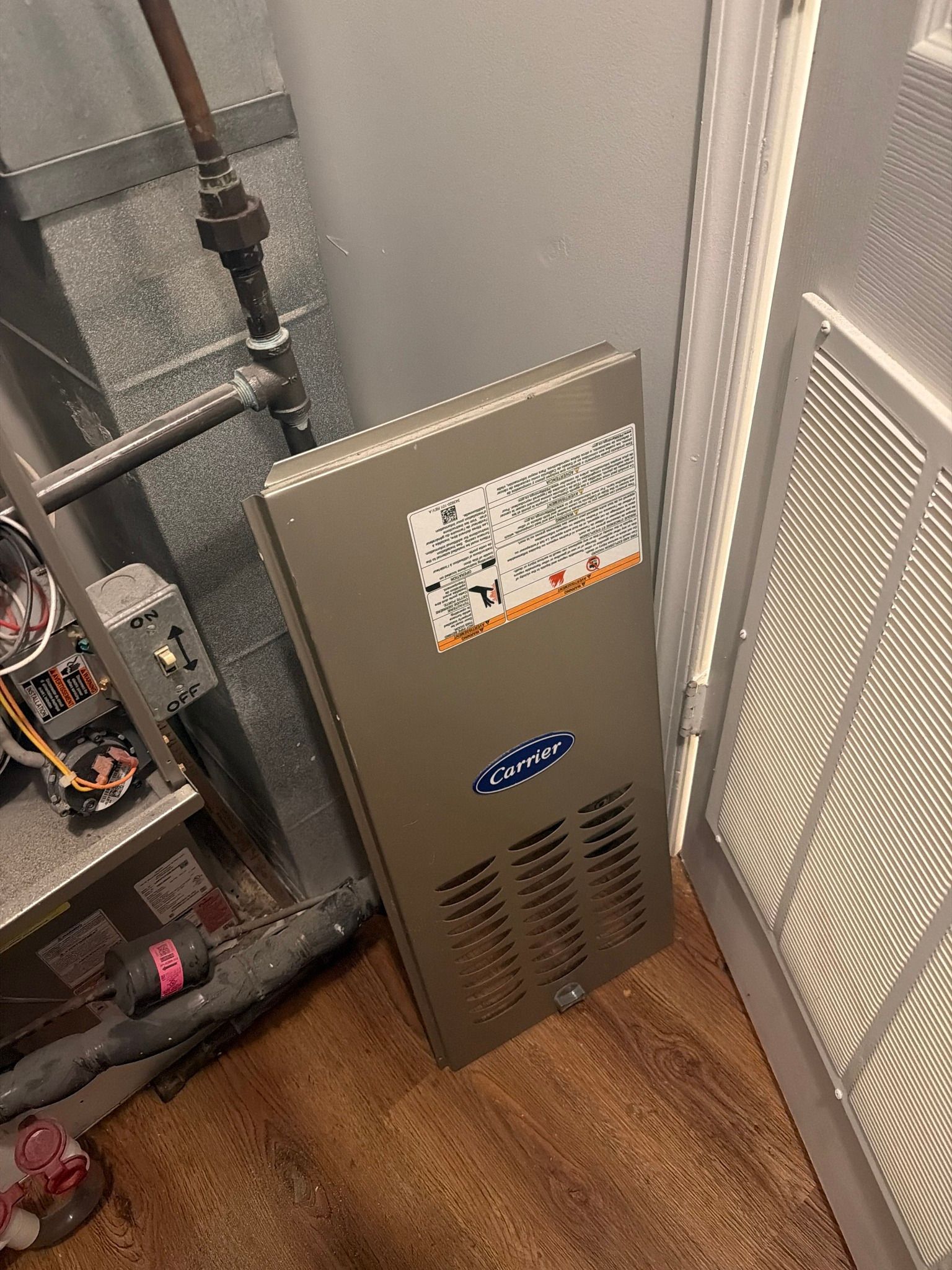 A gray, rectangular furnace panel with a Carrier logo leaning on the floor next to an HVAC unit and a return air vent.