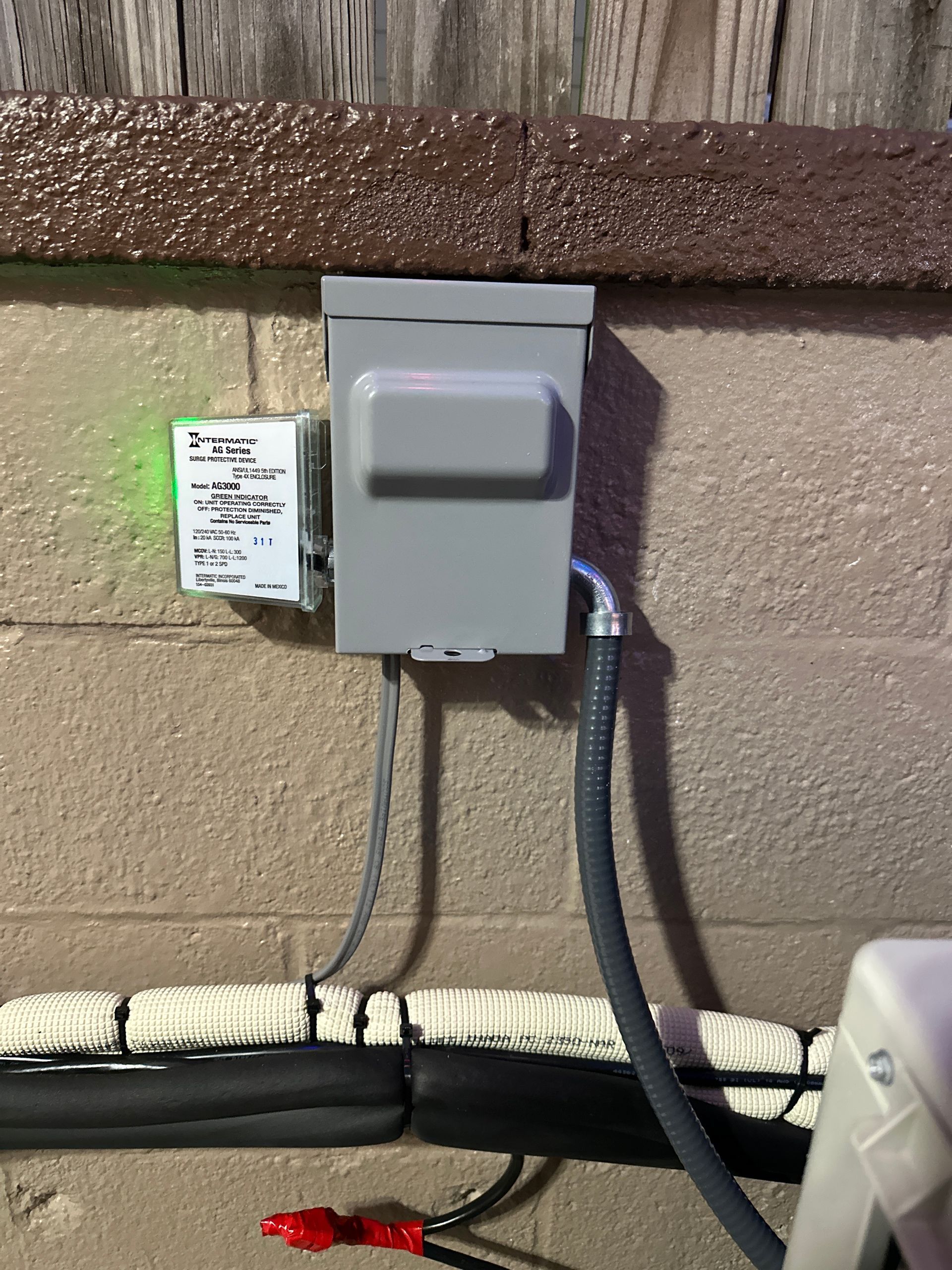 A gray electrical disconnect box and a small white device with a green indicator light mounted on a concrete block wall.