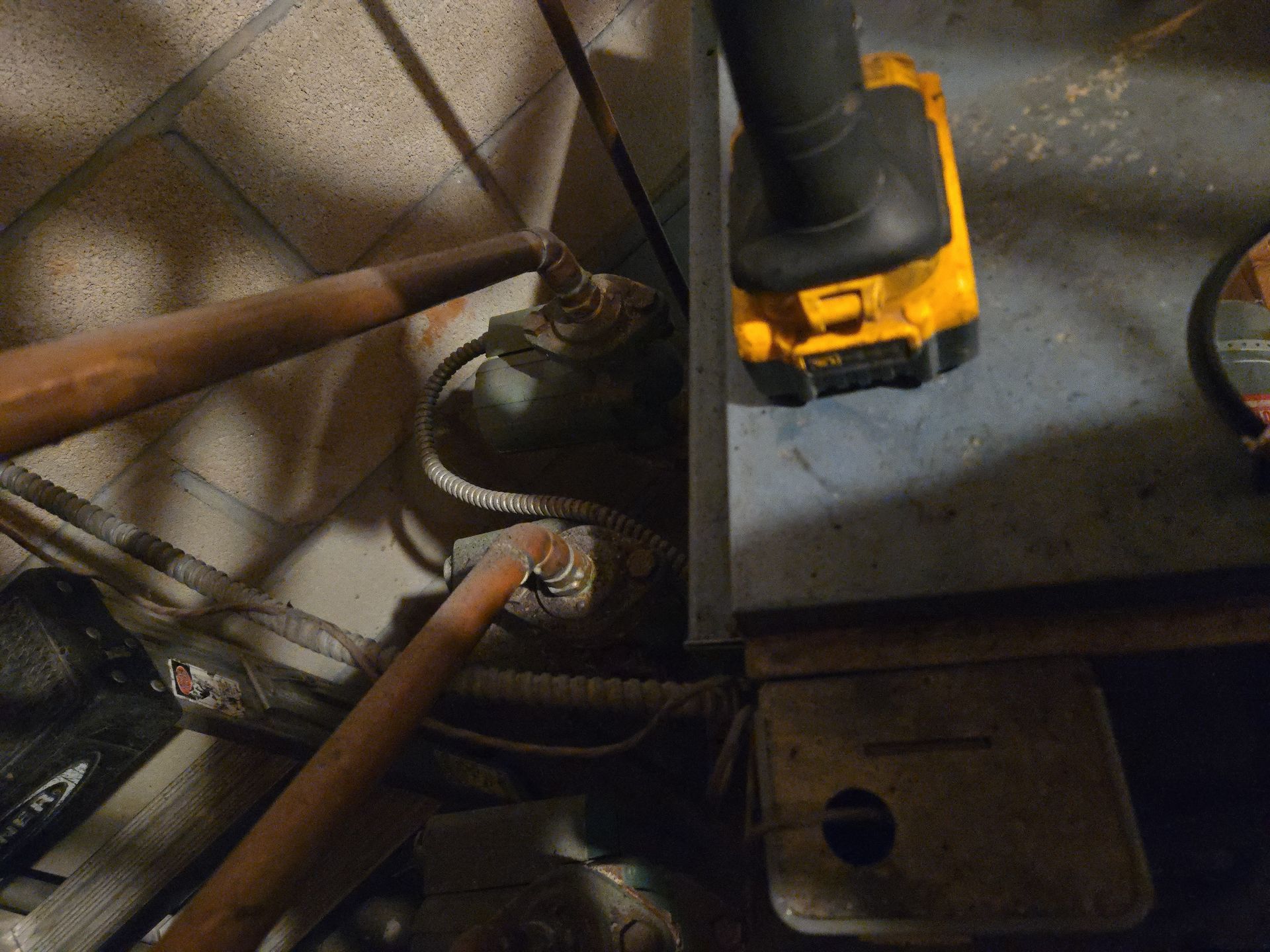 A yellow power tool battery sits atop a dark metal surface near two copper pipes connected to equipment in a utility area.
