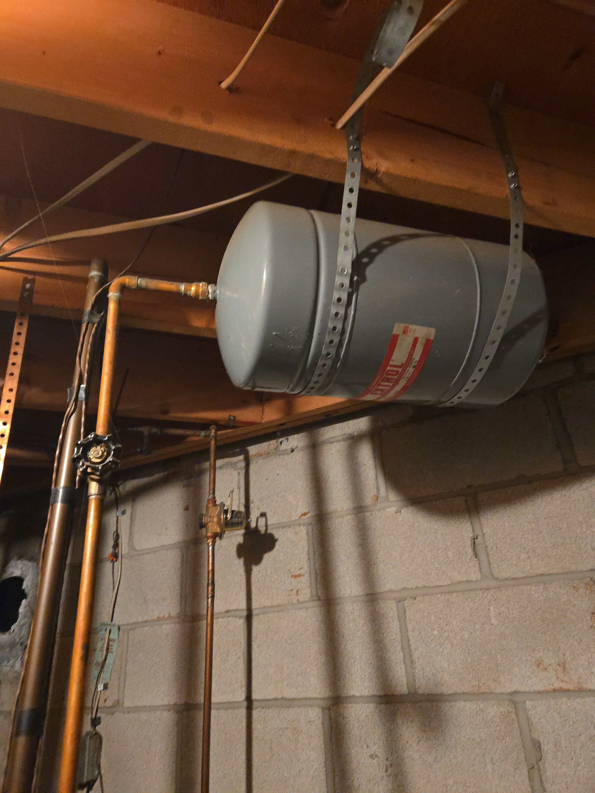 A grey water expansion tank hanging from wooden ceiling joists in a basement, connected to vertical copper plumbing.