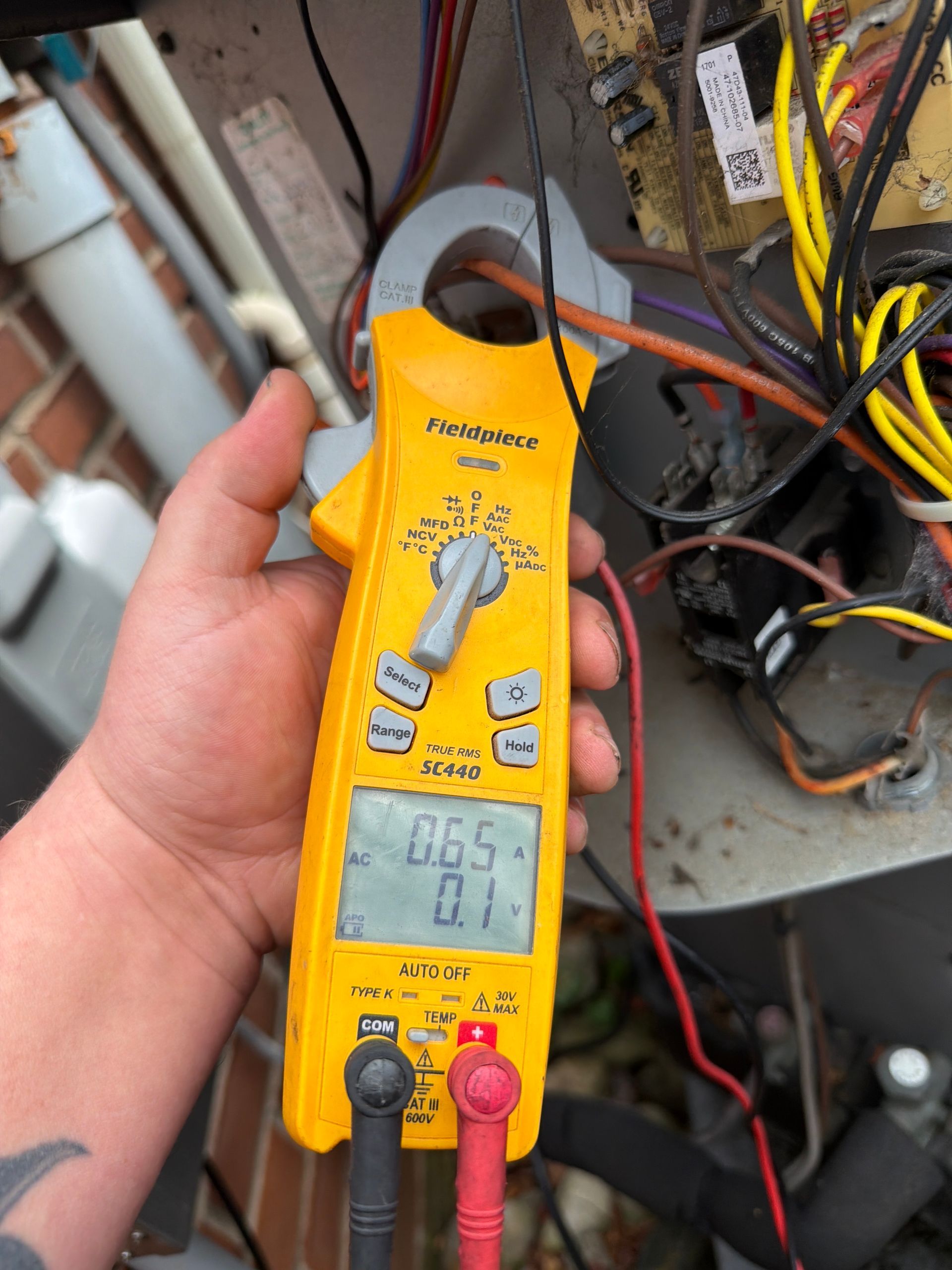 A hand holding a yellow digital multimeter measuring electrical current on a wire inside an open HVAC unit.
