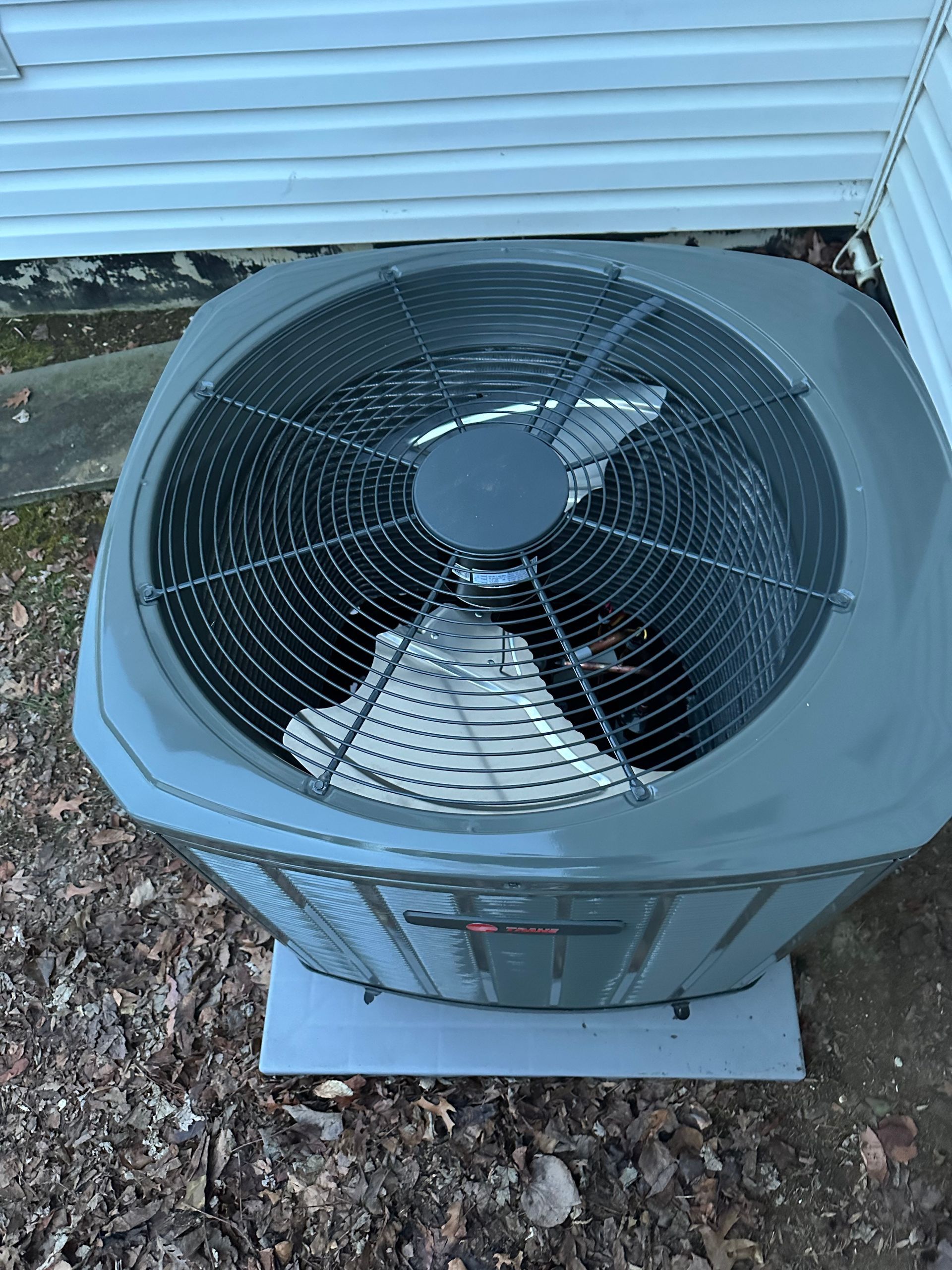A gray outdoor air conditioning condenser unit sits on a concrete pad against white siding, viewed from above.