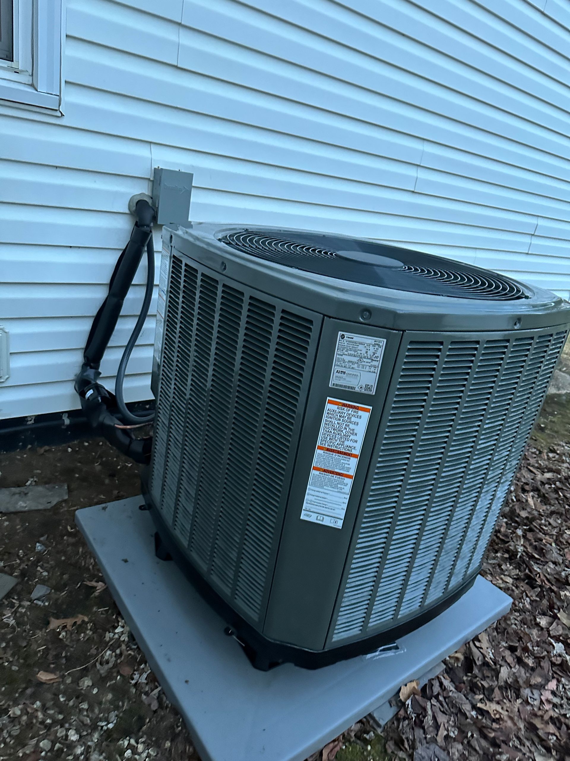 A grey, square-shaped outdoor central air conditioning unit sits on a concrete pad against a white-sided house wall.