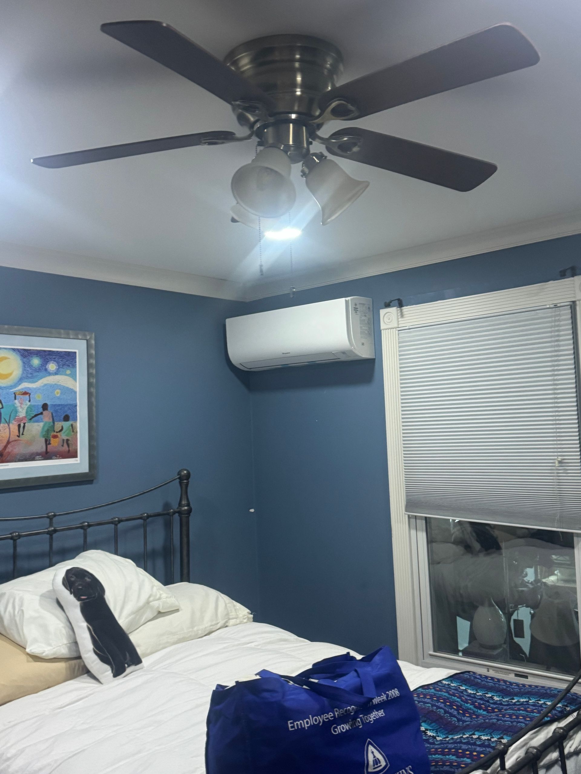 A bedroom with blue walls, a ceiling fan, a wall-mounted AC unit, a bed with pillows, and a window with blinds.