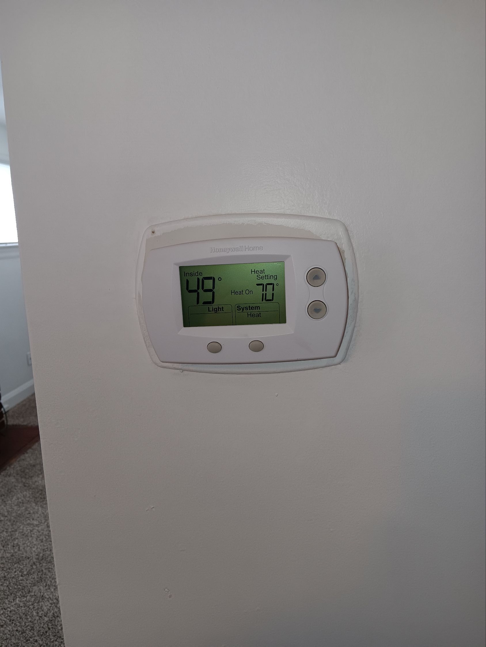 A white digital thermostat mounted on a plain wall, displaying a room temperature of 49 degrees.