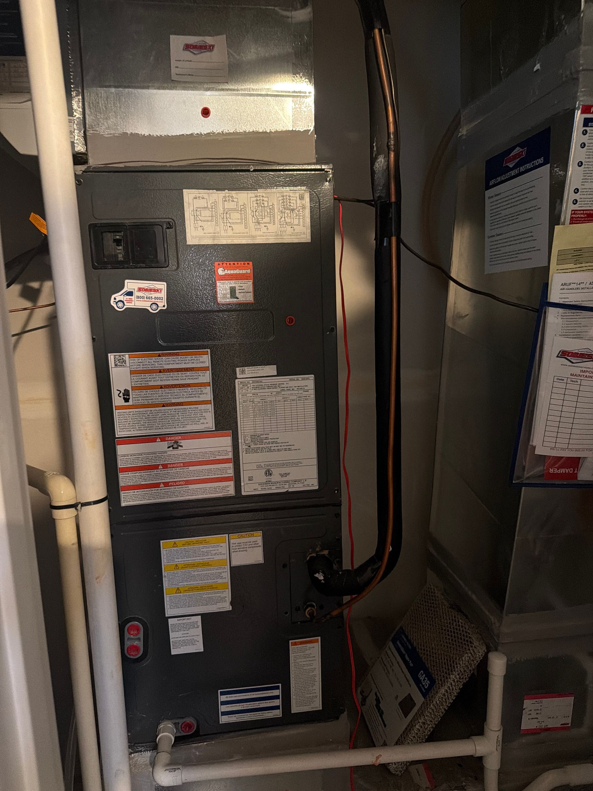 A dark gray HVAC furnace unit with several warning labels stands upright next to metal ductwork and white PVC pipes.