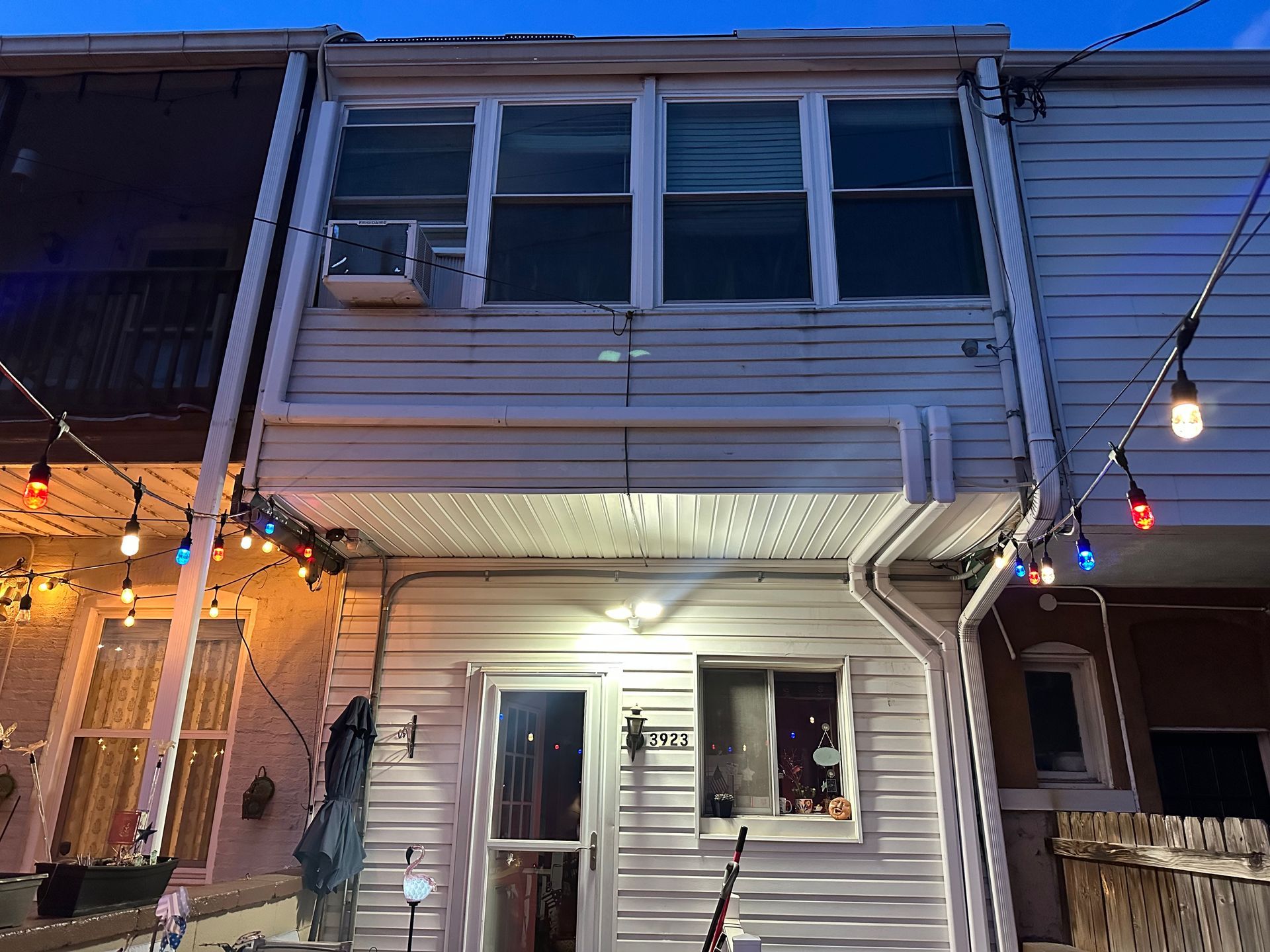 A two-story row house at dusk with light-colored siding, a small balcony window, and string lights hanging in front.