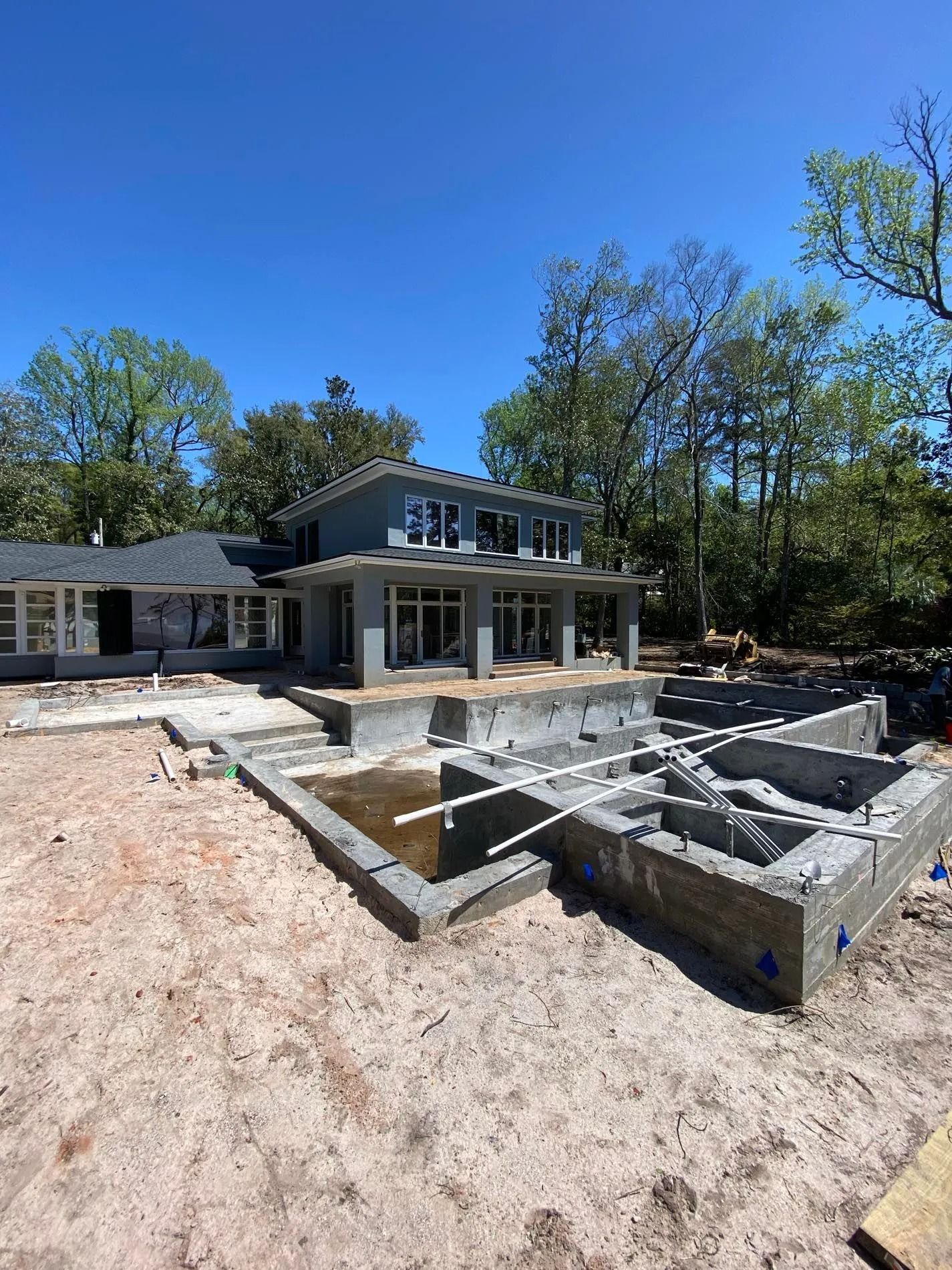 Construction site with unfinished house and pool, blue sky, surrounded by trees.