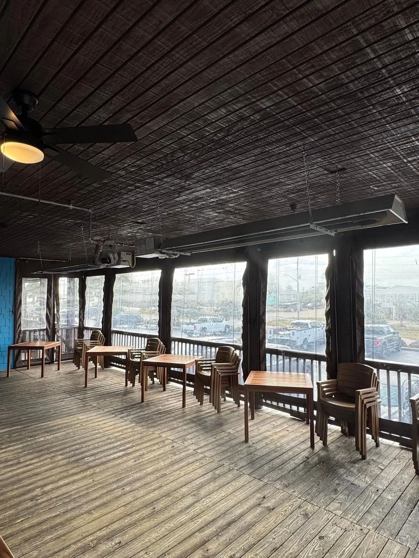 Outdoor dining area with wooden tables and chairs, large windows, and a dark wooden ceiling.