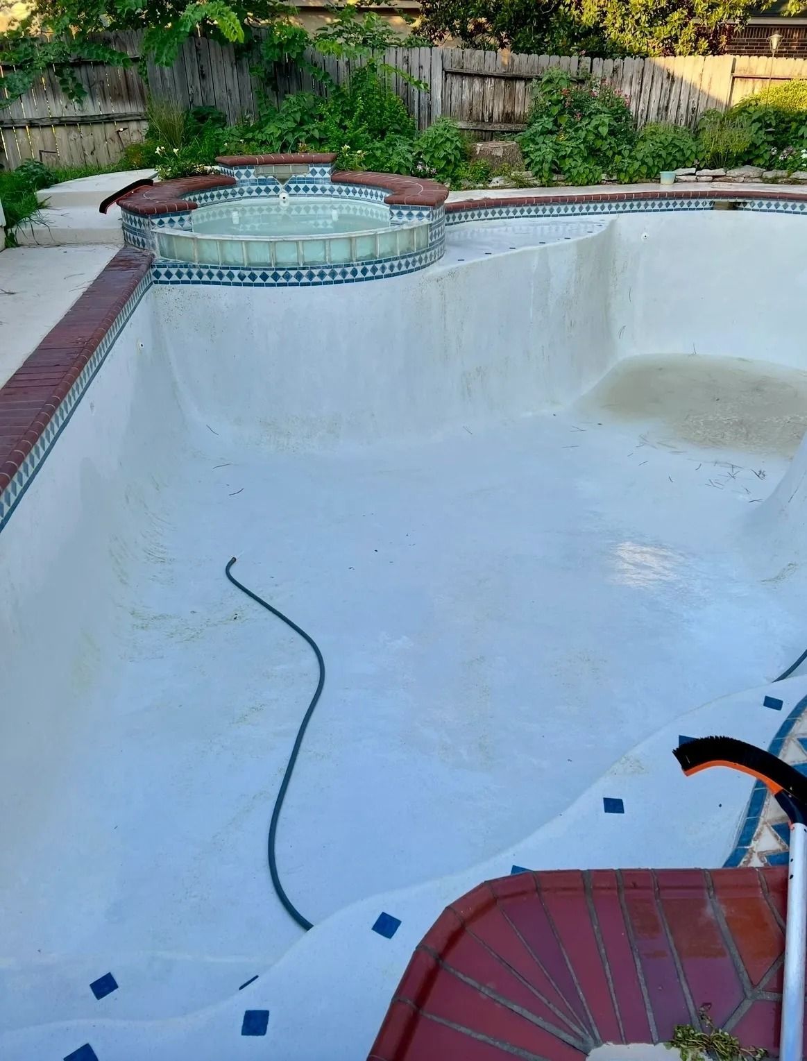 Empty white swimming pool with a hose inside and a tiled spa at the edge, in a backyard setting