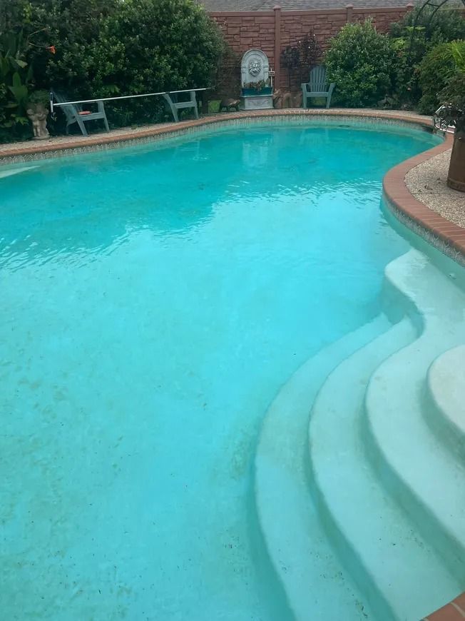 Curved backyard swimming pool with turquoise water, white steps, and brick patio seating in the background
