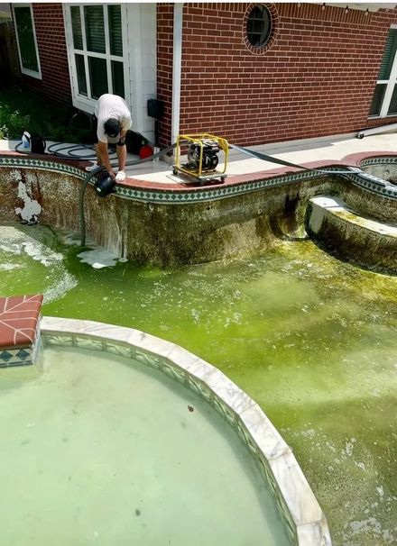 Worker cleaning a green backyard pool beside a brick house and patio equipment.