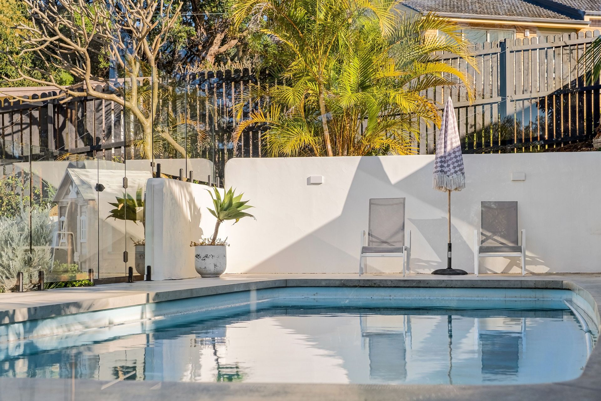 Backyard pool with two lounge chairs, umbrella, potted plant, and white fence in sunlight