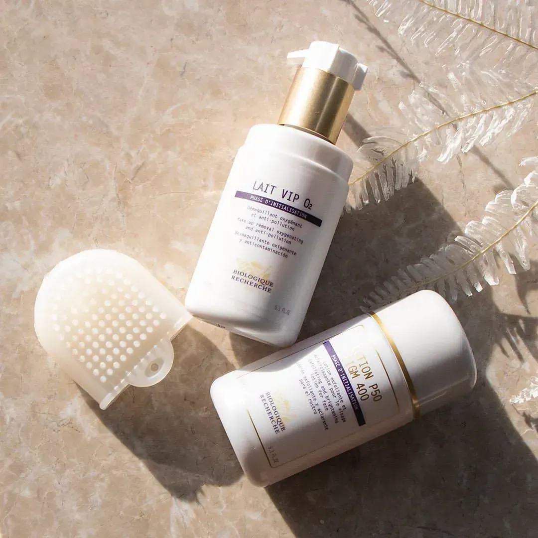 Two white skincare bottles with gold accents and a silicone face scrubber on a light surface.