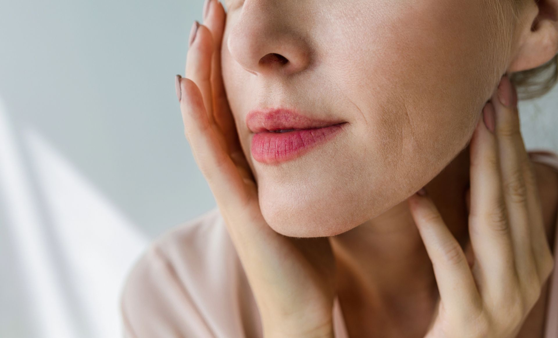 Woman's face with hands gently touching cheeks and neck; lips are pink.