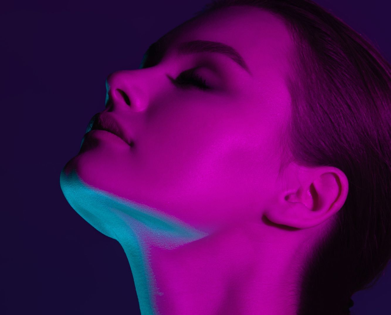Woman's profile lit with vibrant magenta and teal. Eyes closed, looking up, with a neutral expression. Dark background.