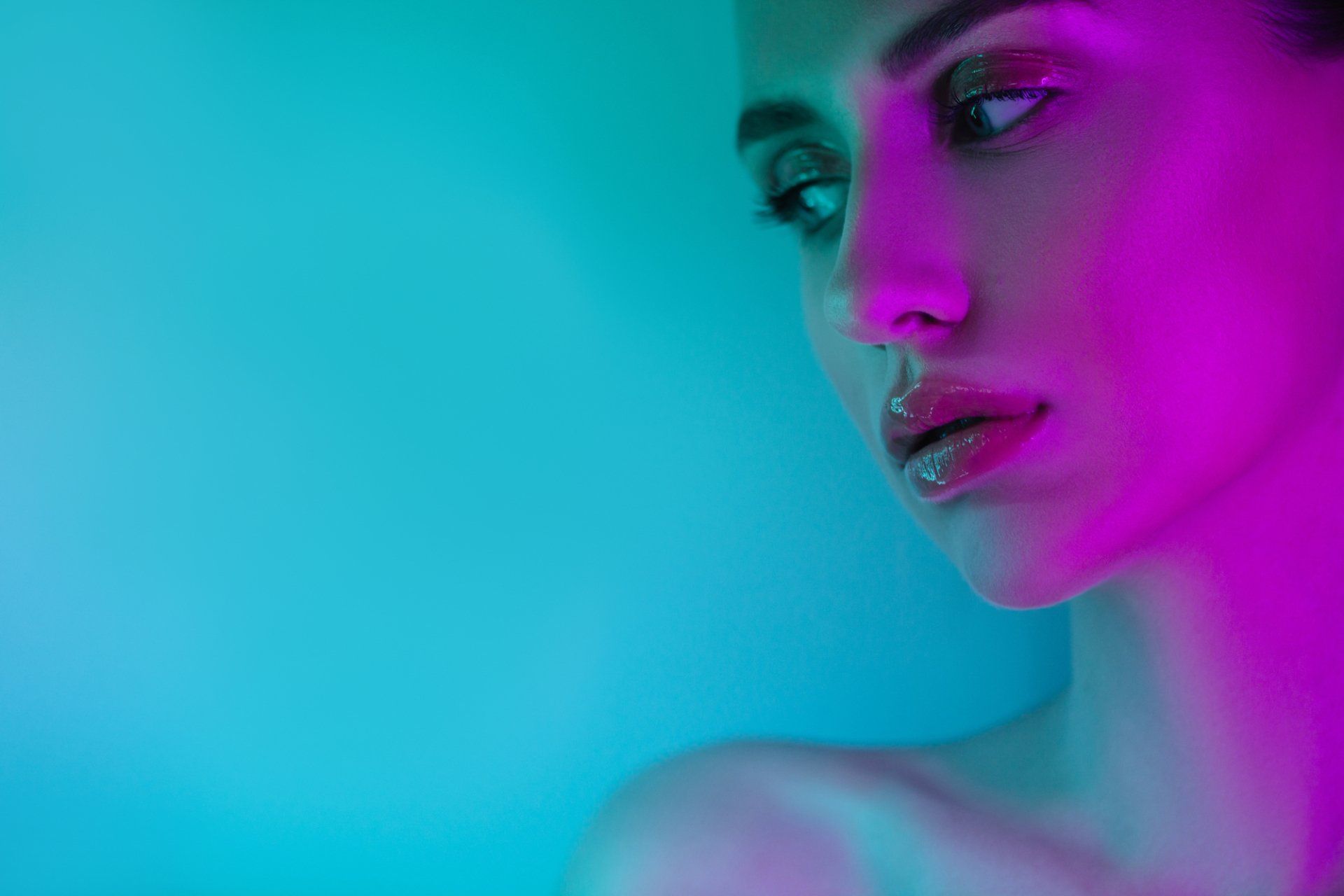 Woman's face illuminated by blue and pink light, looking off-camera with glossy lips.