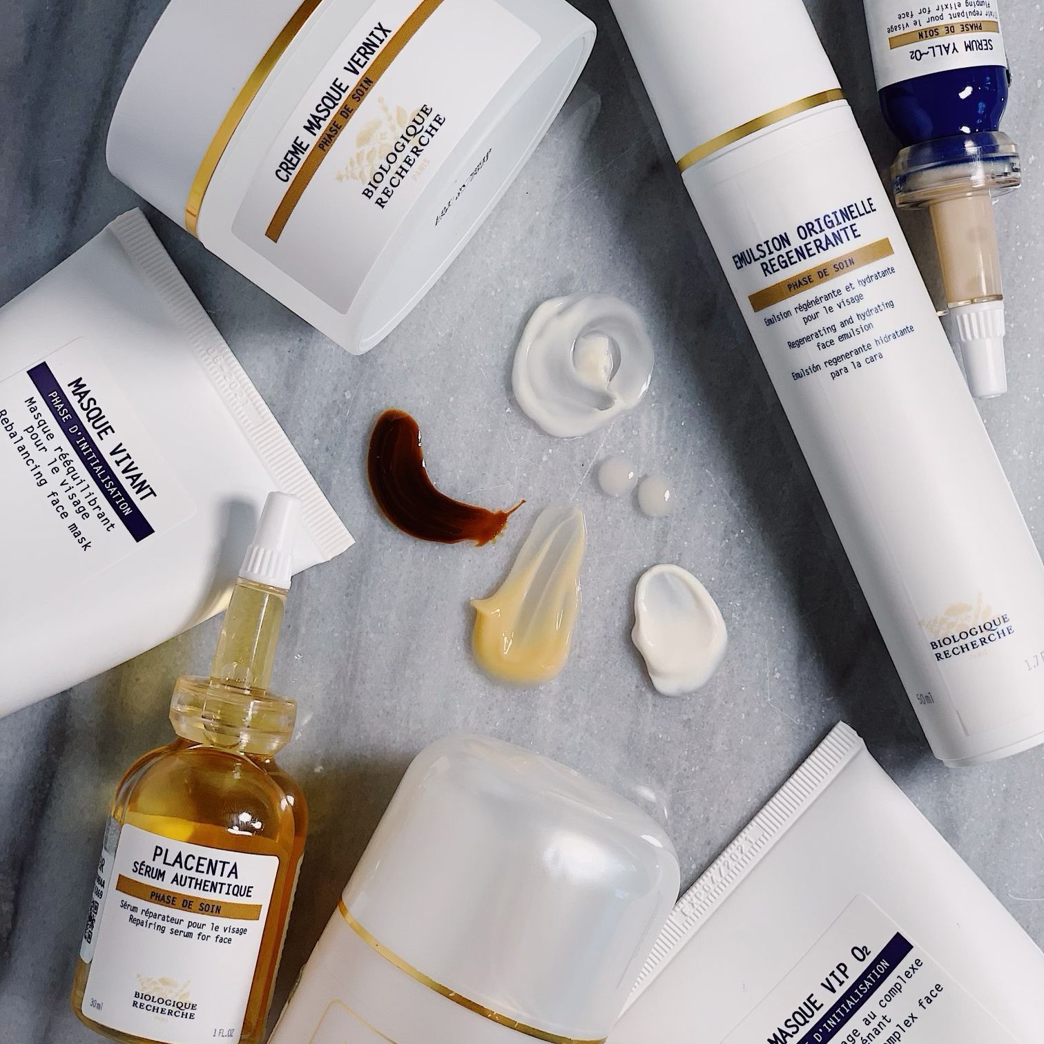 Skincare products, white with gold accents, arranged with different colored creams and serums, on a light gray surface.