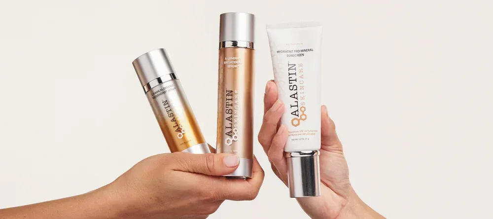 Hands holding three Alastin skincare product tubes with silver and gold tones.