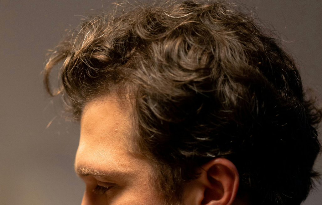 Close-up of a person's head, focused on brown, wavy hair and ear, with a muted background.