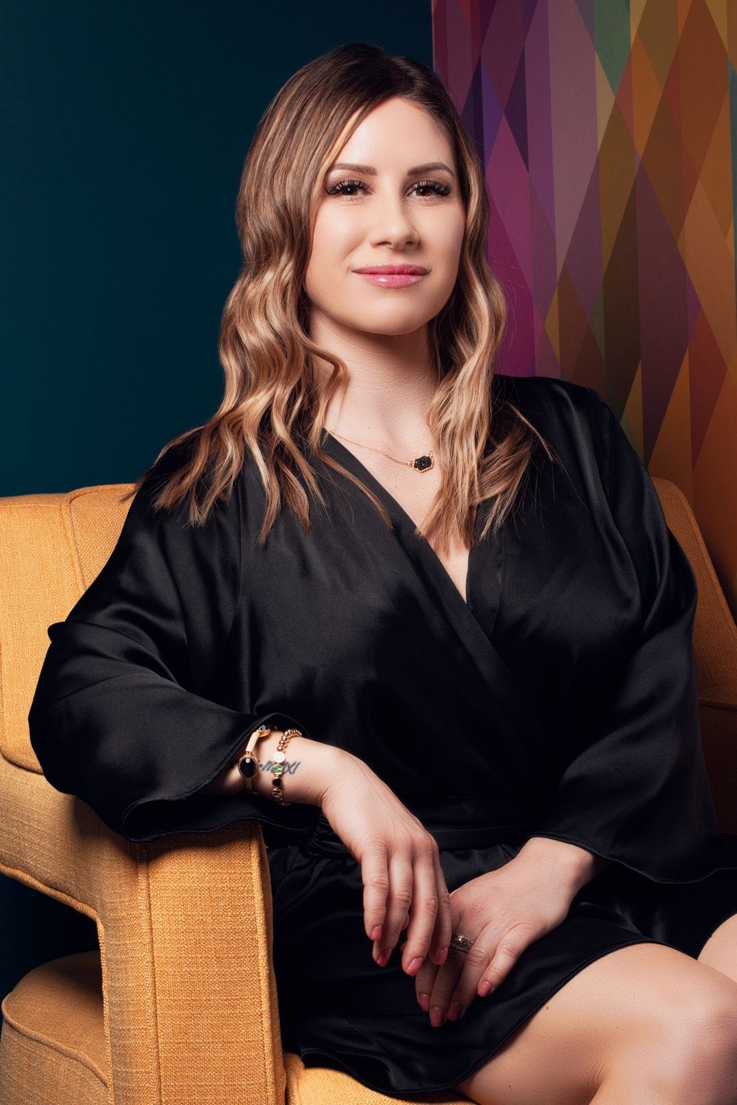 Amanda O’Neill, Licensed Esthetician in black dress sits in a yellow armchair, smiling. Colorful geometric pattern background.