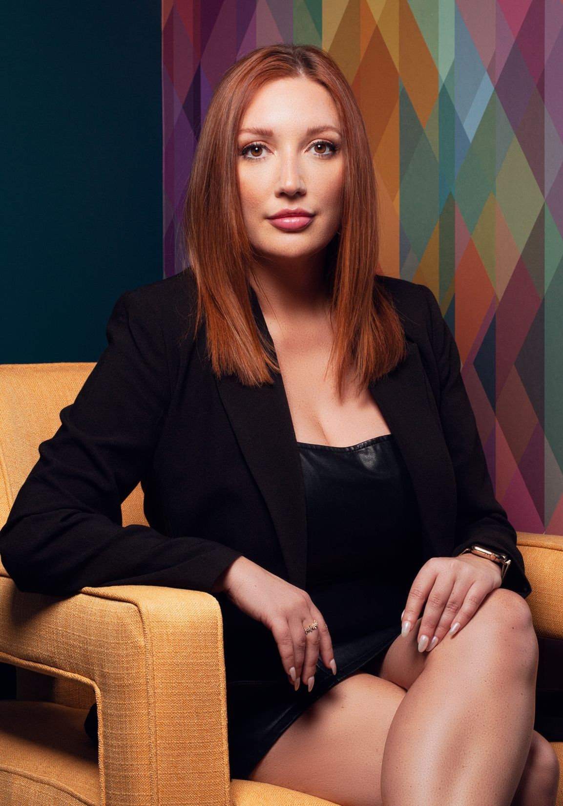 Ashley Knott, Esthetician, sits in a yellow chair, wearing a black blazer and dress, and looking at the camera.