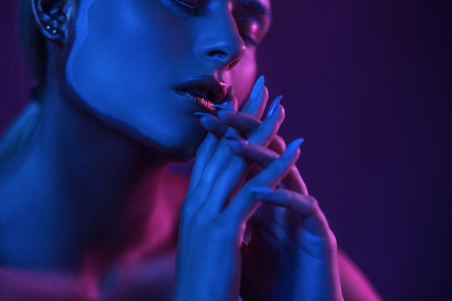 Woman's face and hands illuminated by blue and pink neon light; hands near lips.