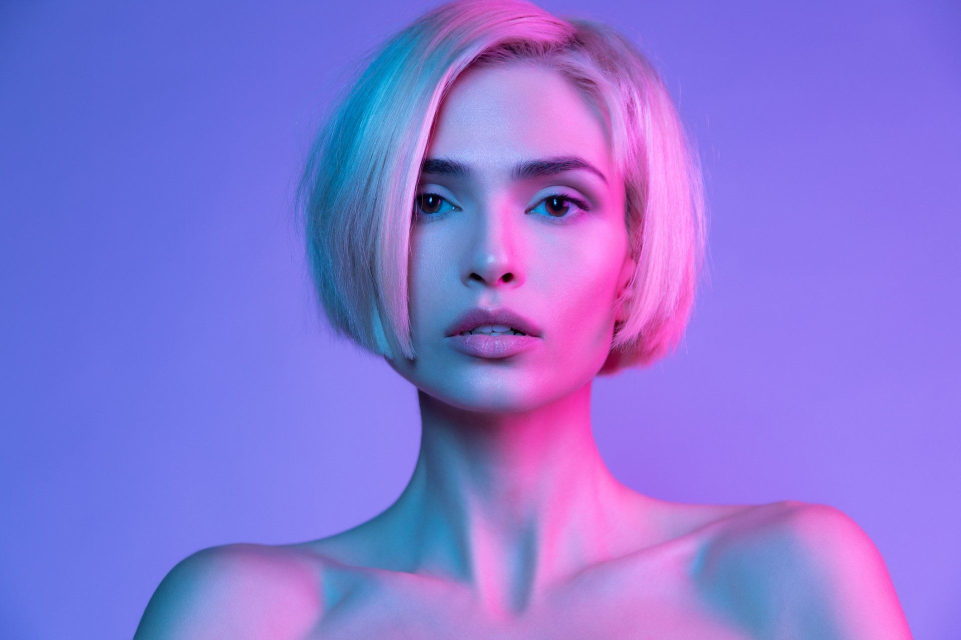 Woman with short blonde hair, lit with pink and blue light, looking directly at the viewer.