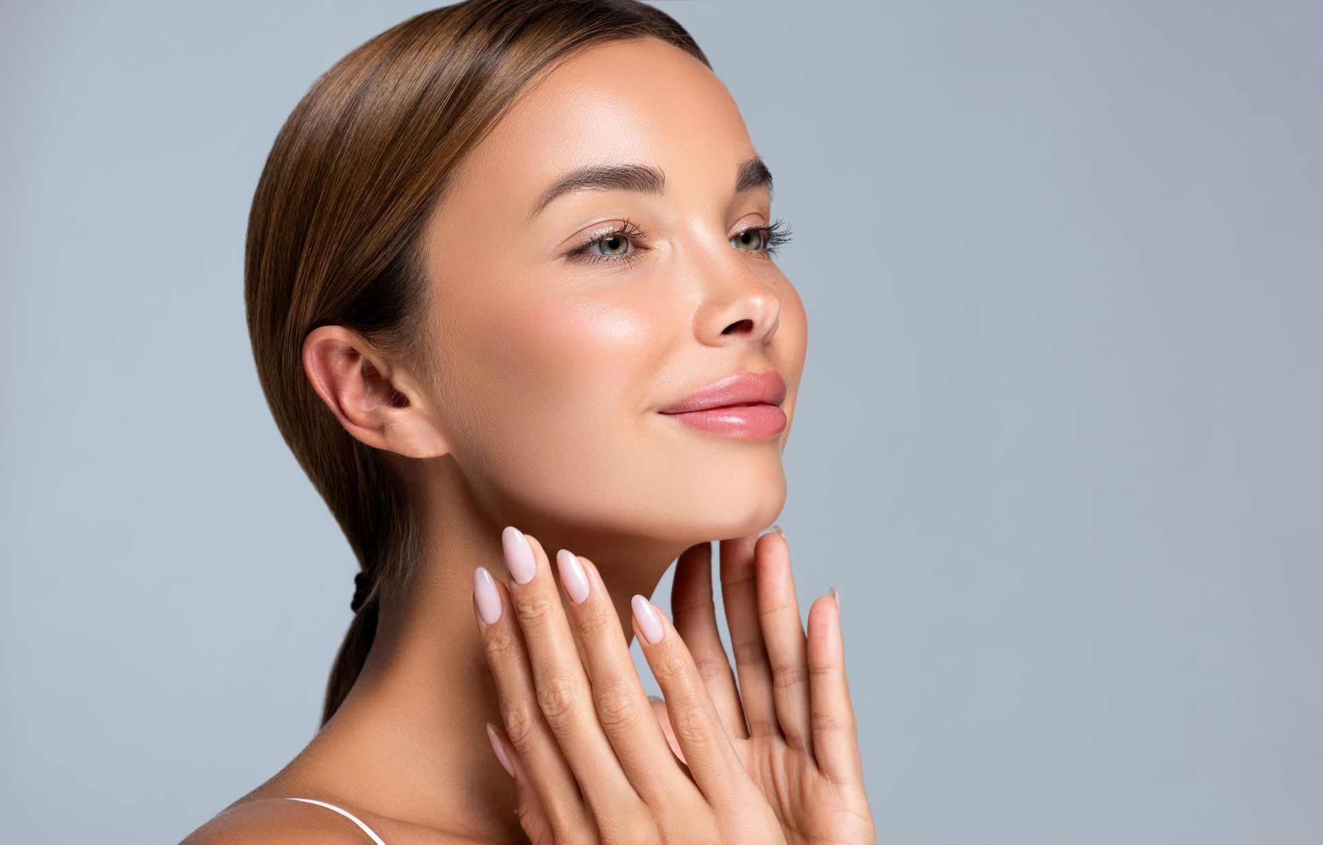 Woman with smooth, glowing skin, hands near face, looking upwards with a gentle smile against a gray backdrop.