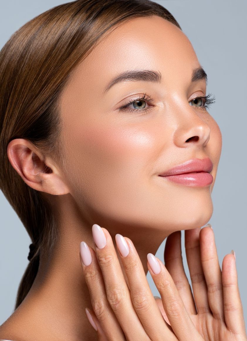 Woman with light skin, hands under chin, eyes closed, rosy cheeks, light pink nails, and a light gray background.