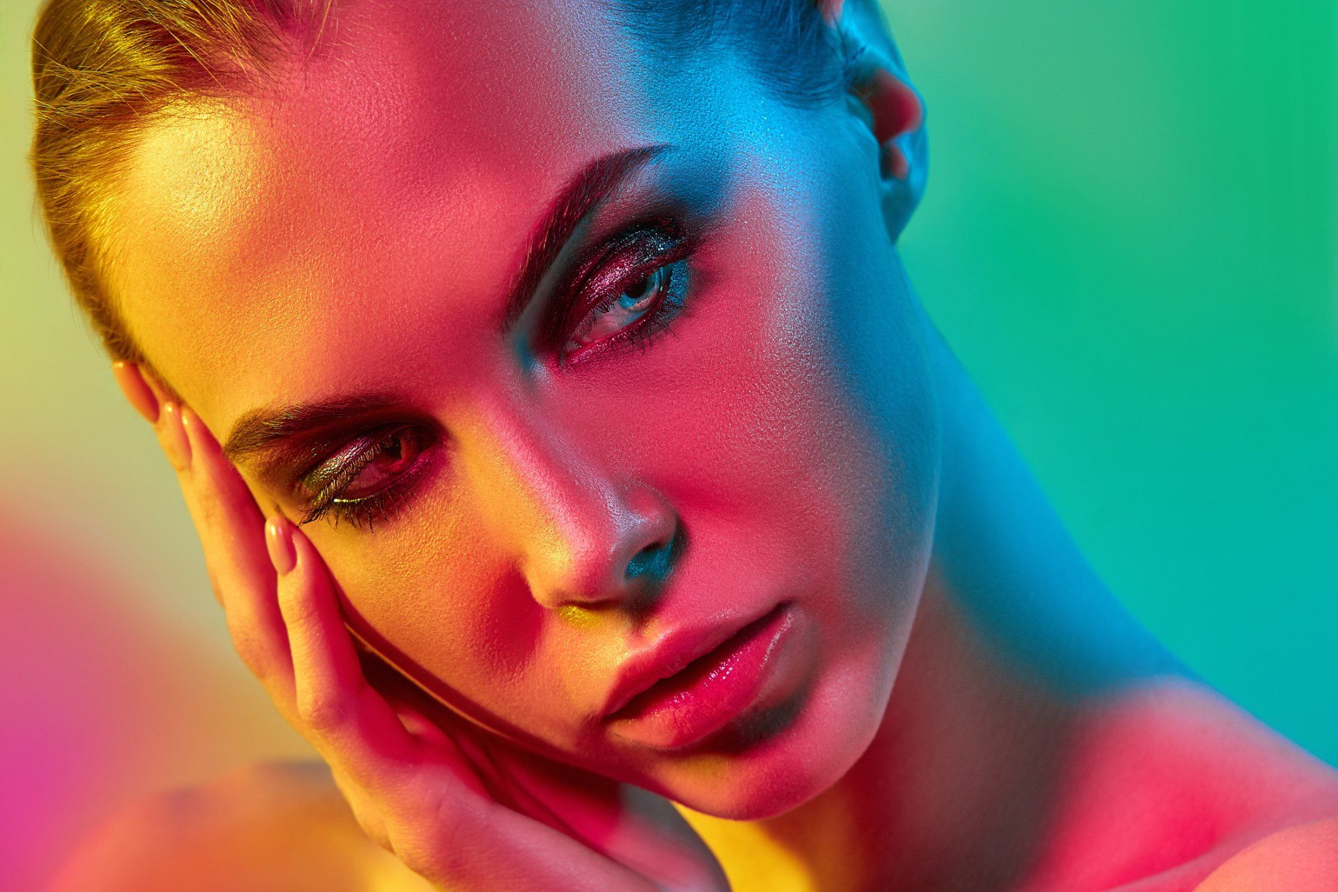Woman's face illuminated by vibrant, multicolored lights. She has bold eye makeup and touches her cheek.