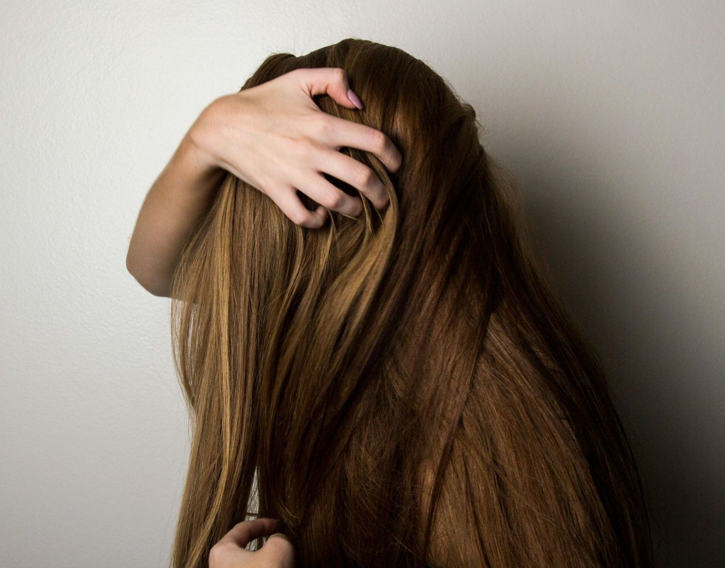 Person scratching head, long brown hair, against a neutral wall.