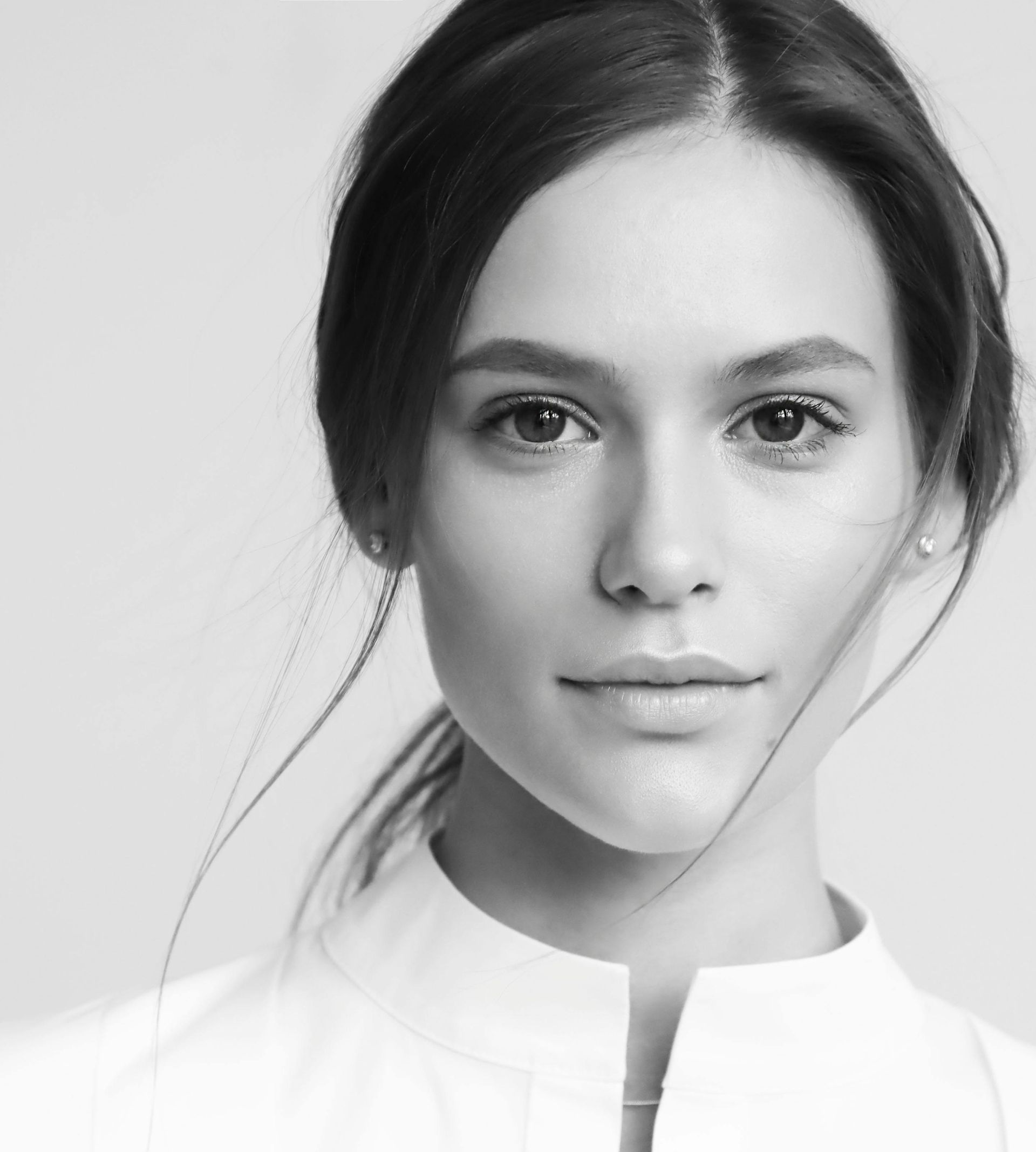 Woman with dark hair pulled back, wearing a white collared shirt, looking directly at the viewer with a neutral expression.