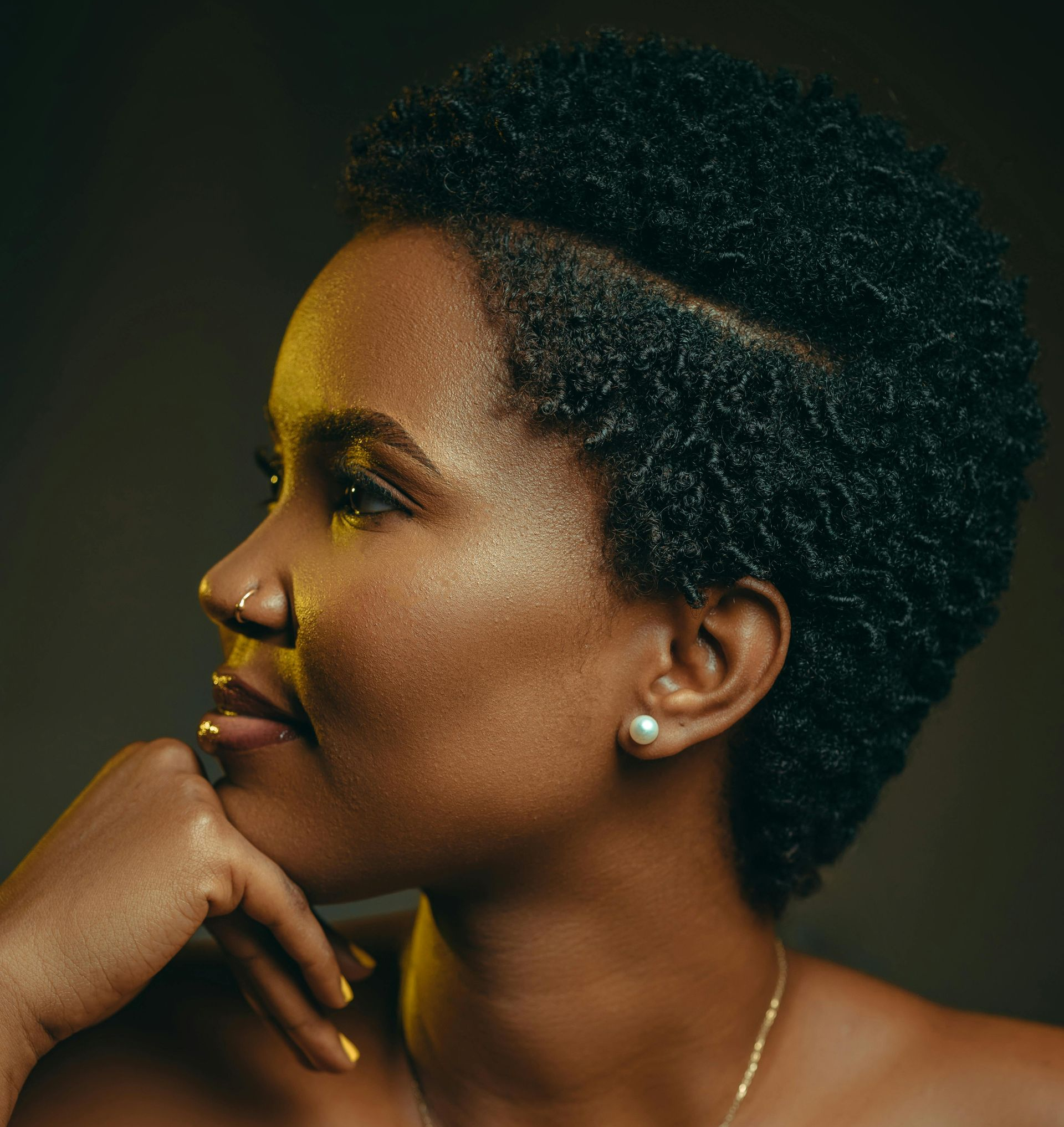 Woman with short, textured hair gazes right, hand on chin. Gold makeup. Neutral background.