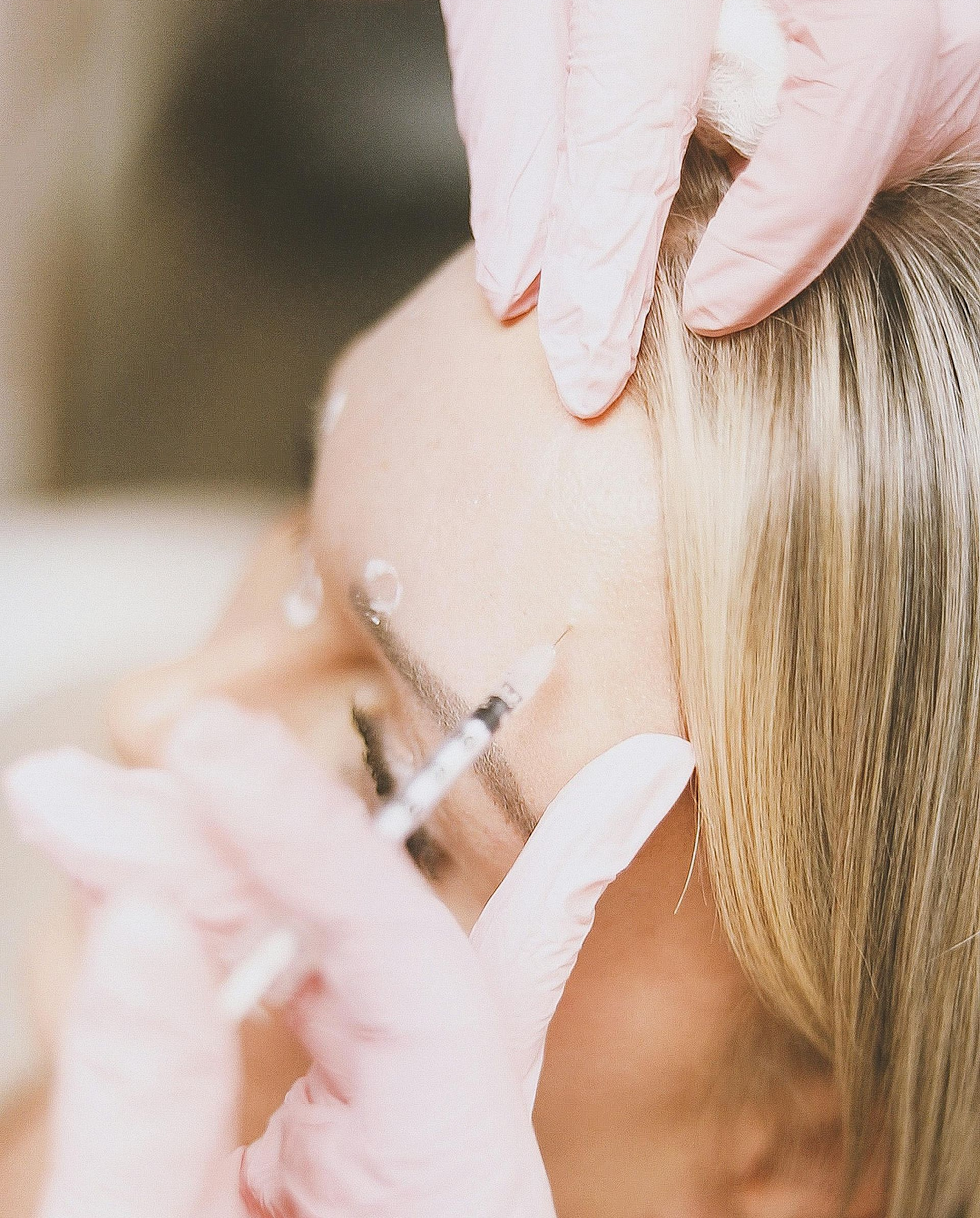Person receiving an injection in the face. Gloved hands hold a syringe near the skin. Blonde hair visible.