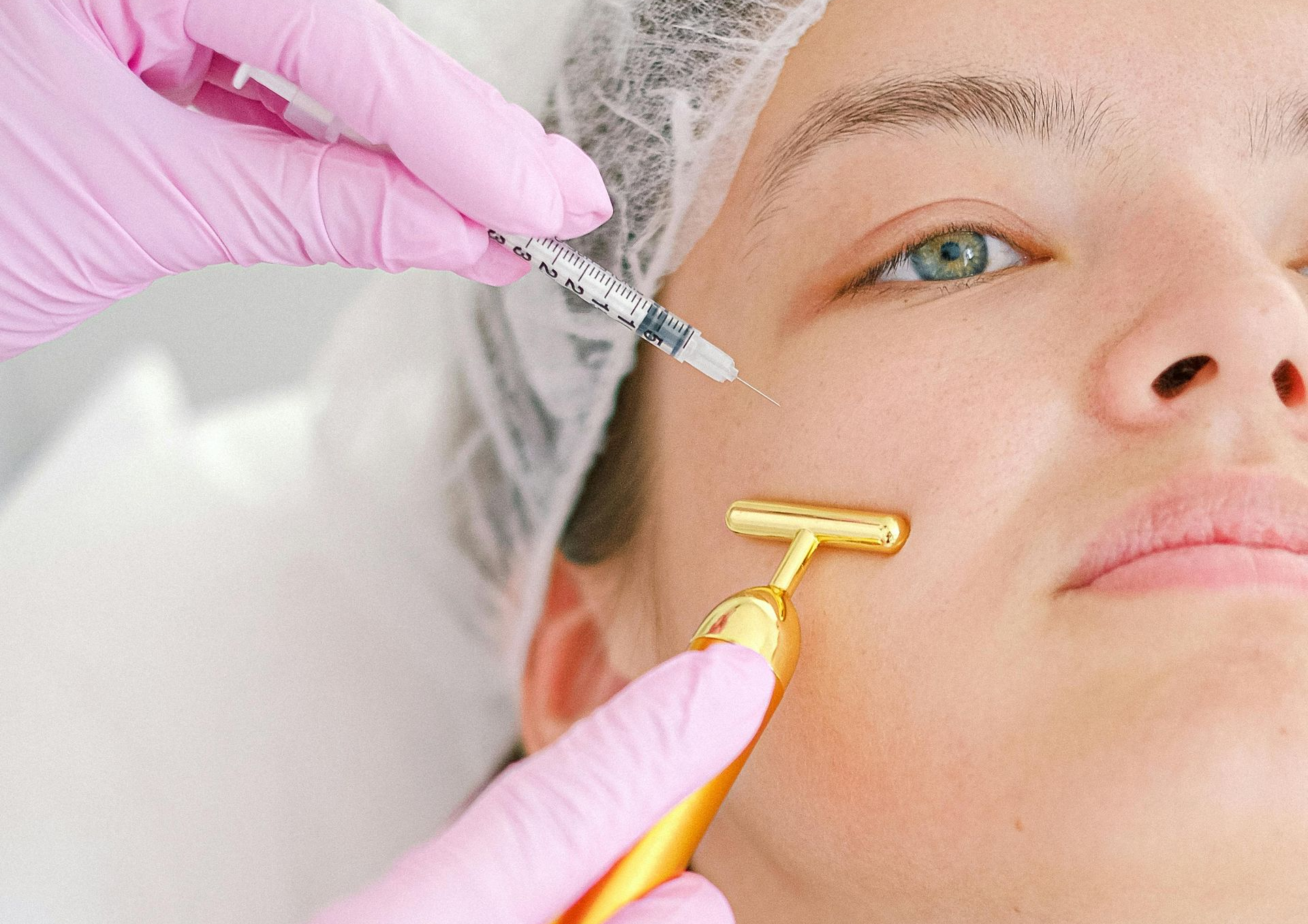 Person receiving cosmetic treatment: needle injection and face roller application, pink gloves.