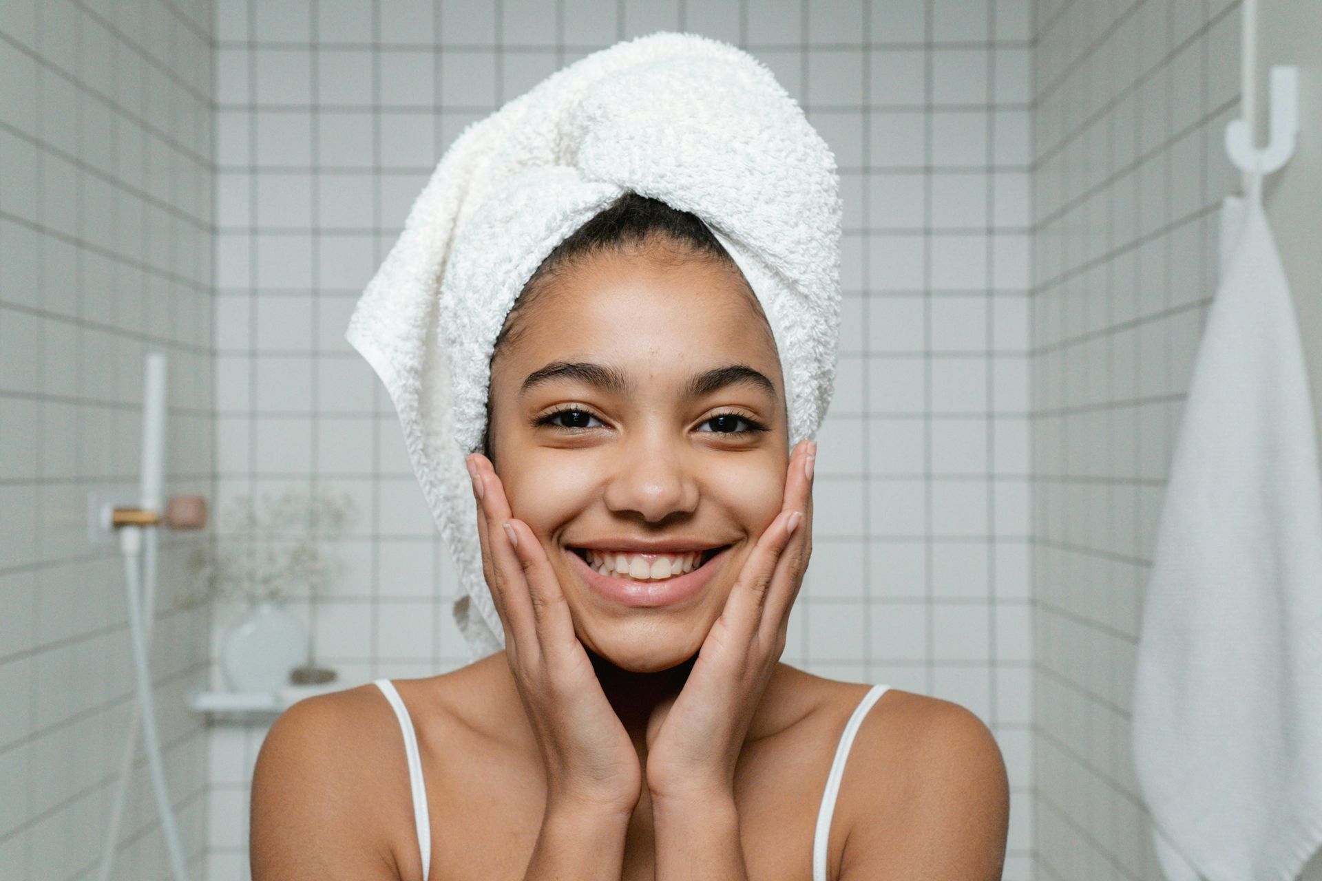 Woman with towel on head smiles, touching cheeks in bathroom.