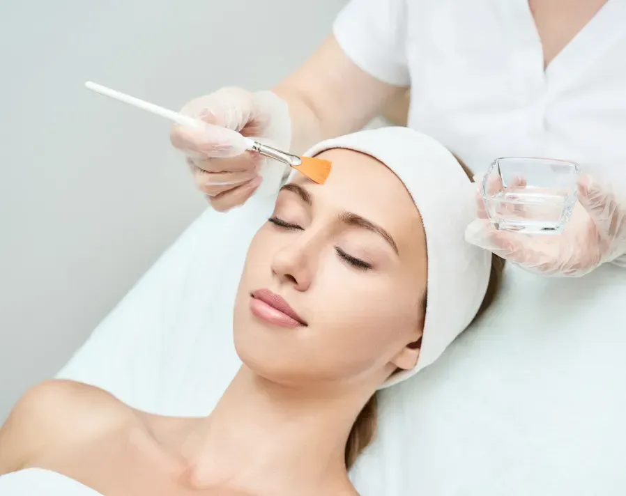 Woman receiving a facial peel, lying down with eyes closed. Aesthetican applies liquid with brush.