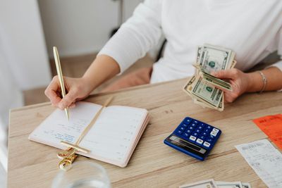 A woman is sitting at a table counting money and writing in a notebook.