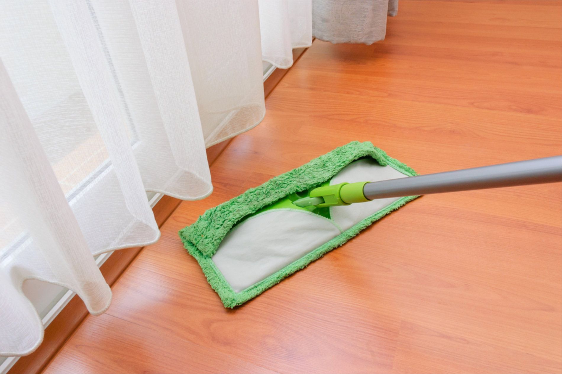 Green and white flat mop cleaning a wooden floor near a window with white curtains — RWM Cleaning in Deception Bay, QLD
