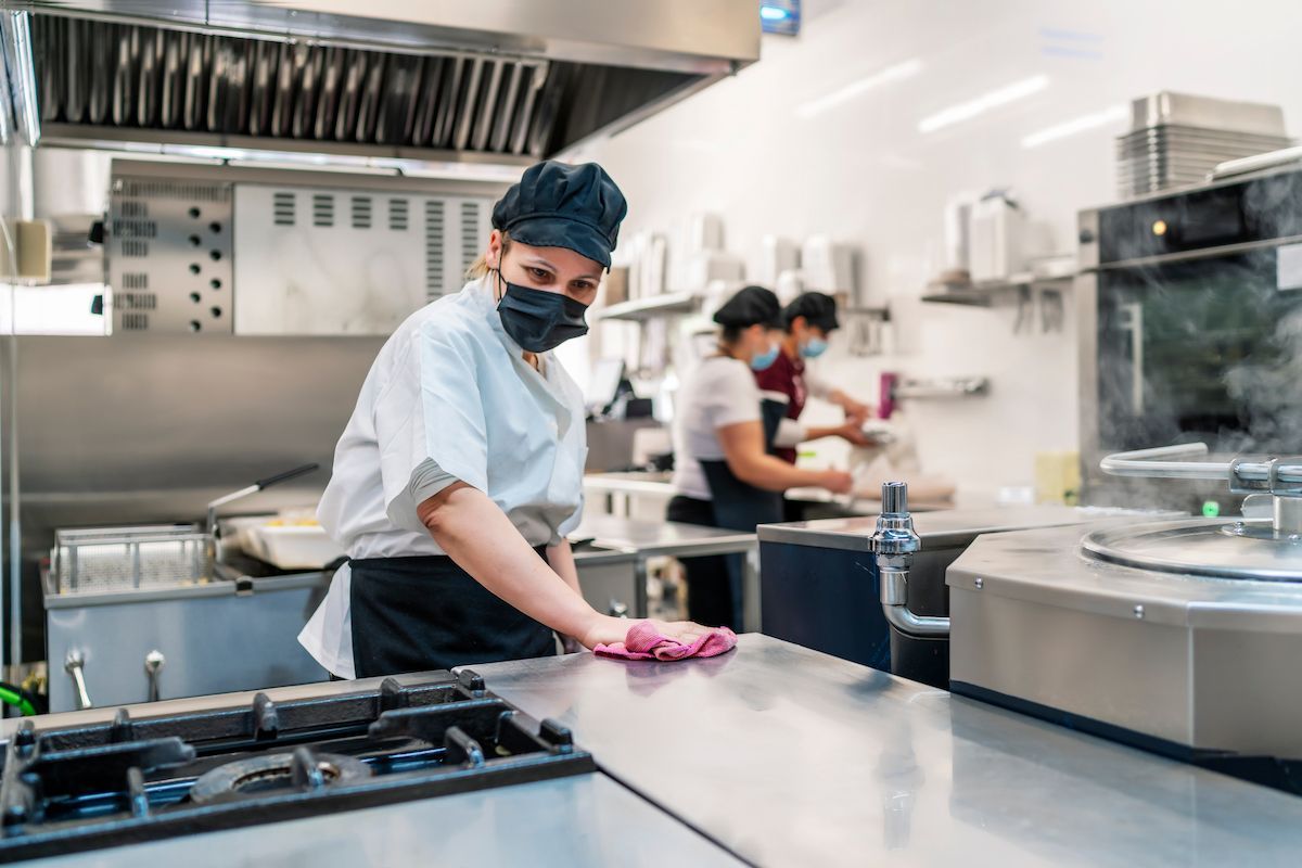 Chef wiping stainless steel counter in commercial kitchen, others preparing food — RWM Cleaning in Deception Bay, QLD